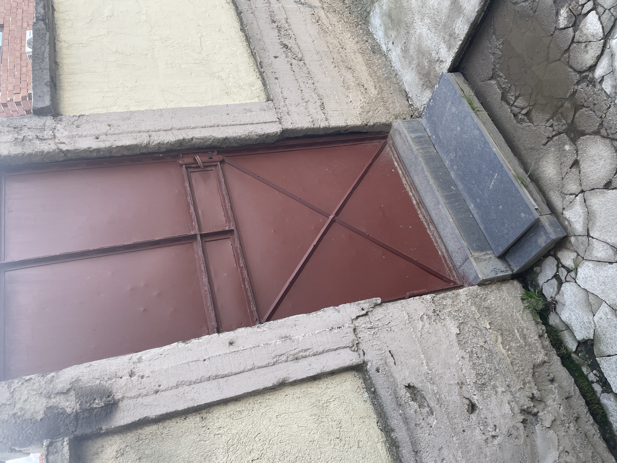 This image captures an angled, overhead view of a weathered outdoor scene in Ihtiman, Bulgaria. The primary subject is a dark reddish-brown metal door or hatch, likely providing access to a basement or utility space. The door is rectangular, featuring a grid of raised seams that divide its surface into panels, and exhibits minor dents and general wear. A simple metal latch mechanism is visible on its upper left side.
The metal door is set within a robust frame of rough, grey concrete structures that display considerable aging, including cracks, crumbling edges, and general wear. Above the door, to the left, a section of a light yellow, textured building wall is visible. Further to the upper left, a small portion of a red brick wall can be seen, along with what appears to be an air conditioning unit.
To the immediate right of the metal door, a small set of dark grey or black steps, likely composed of stone or concrete slabs, ascends slightly. Beyond these steps, the ground transitions into irregularly shaped, cracked concrete or stone paving, characterized by numerous gaps and crevices. Small patches of green grass and moss are growing through these cracks and along the edges, suggesting a neglected or long-established area.
The scene is brightly lit, indicative of daytime, with relatively even illumination across the various surfaces. The textures are prominent, from the coarse concrete and cracked paving to the smoother metal of the door and the textured yellow wall. No human or animal activity is depicted, and there is no visible text in the image. The overall impression is one of an aged, utilitarian, and somewhat neglected urban or semi-urban environment.