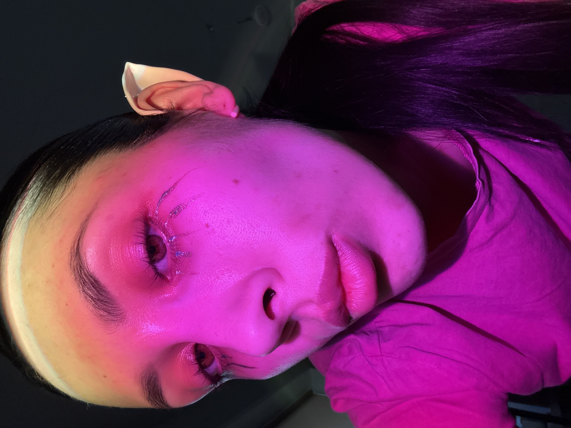 A young person with dark hair is seen close-up, turned at an angle, under pink-purple lighting. A light-colored prosthetic ear is attached near the hairline. Shimmering makeup or adhesive strips adorn the face near the eye. The person's face is resting against a pink garment. No geographical information or any mention of Votkinsk, Russia, is discernible from the image.