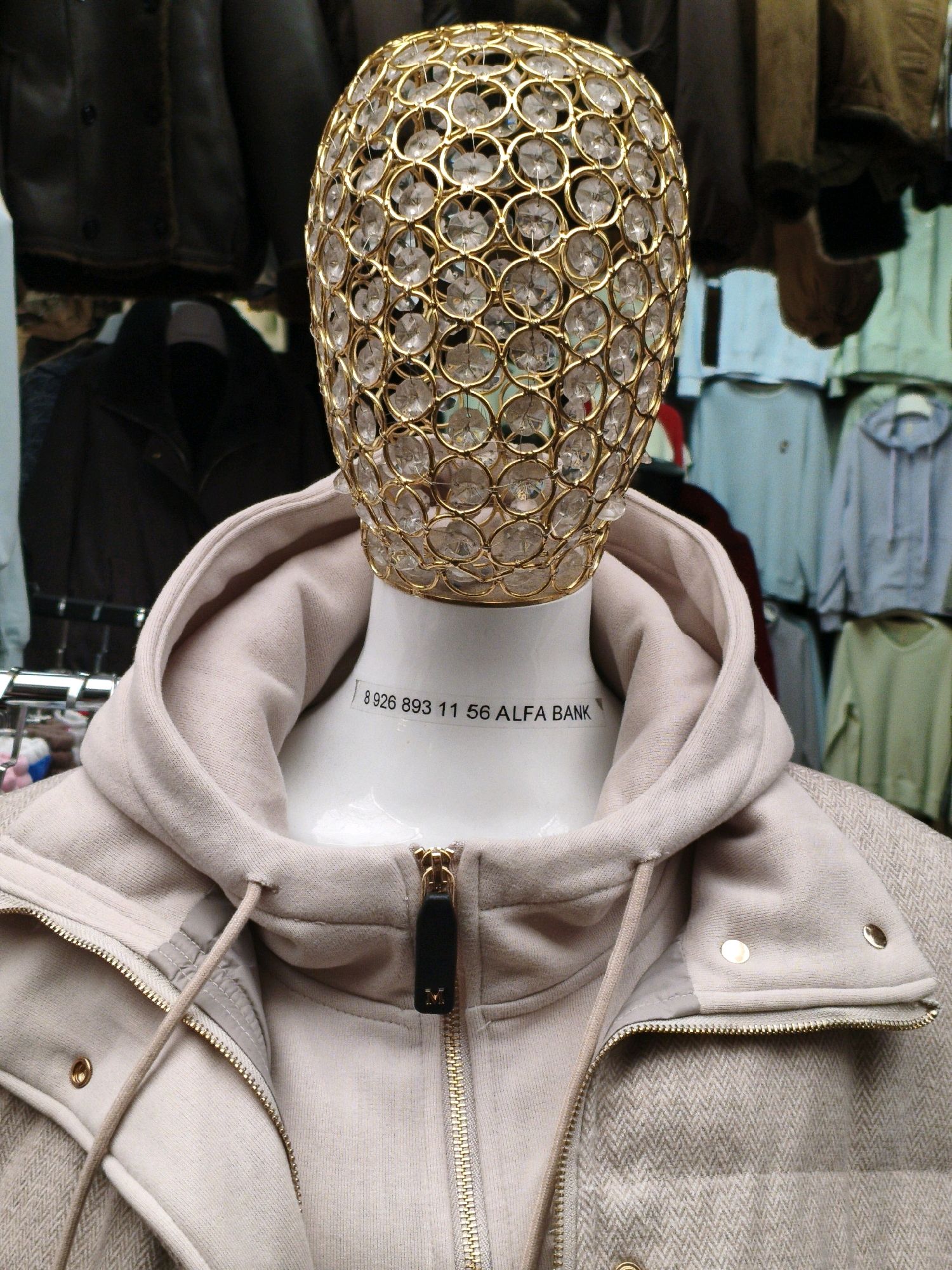 A close-up, eye-level shot shows a mannequin wearing a cream-colored hooded jacket with a herringbone pattern. The mannequin's head is replaced by a gold, cage-like headdress adorned with clear, faceted crystals. A black tag with white text is affixed to the mannequin's neck, reading "8926 893 11 56 ALFA BANK". The jacket has a zipper closure with a gold pull tab and a black leather fob. The background consists of racks of clothing, including several light blue and light green hooded sweatshirts, and dark jackets. The overall impression is of a retail display.