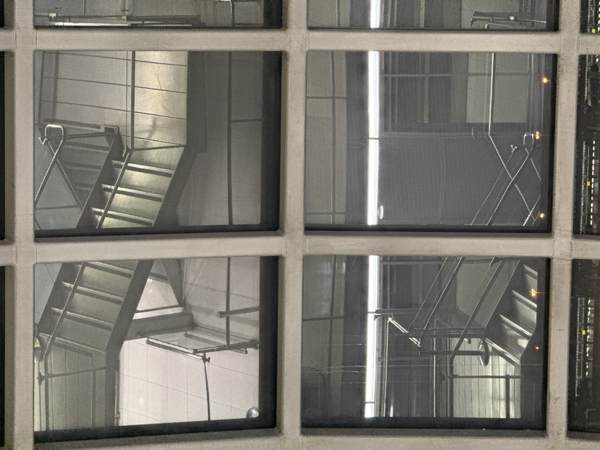 The image captures a view through a grid of windows, likely part of a building's exterior. The windows are separated by concrete frames. Inside, visible through the perforated glass, are multiple levels of modern staircases with metal railings. The staircases appear to be made of metal and concrete, with some sections reflecting light. The overall environment suggests an indoor, possibly public or commercial, space with a focus on architectural design and functionality. The lighting indicates it could be evening or night, given the artificial illumination within. The location is identified as Nong Prue Subdistrict, Thailand. There are no people or discernible actions visible in the scene.