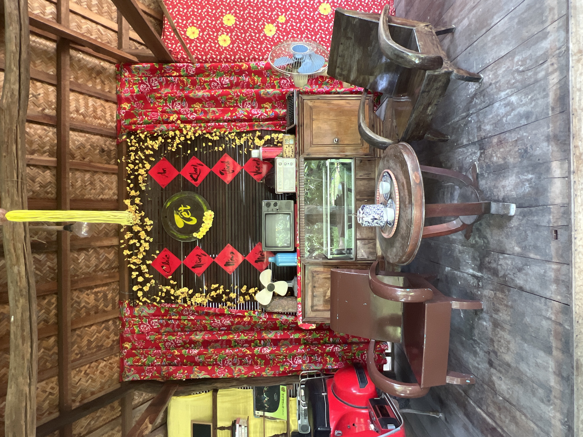 This image captures a corner of a rustic, open-air establishment, likely a cafe or guesthouse, in Hoi An, Vietnam. The scene is decorated for a festive occasion, possibly Tết (Vietnamese Lunar New Year), with vibrant red floral curtains and yellow decorative elements.
In the foreground, a small, round wooden table holds a tea set, consisting of a teapot and two cups arranged on a woven tray. Two vintage wooden armchairs flank the table, one a rich brown and the other a darker, weathered wood, both facing the central display.
The background is dominated by a wooden cabinet topped with various items. A retro television set sits atop the cabinet, beside a vintage radio and a blue ceramic bottle. A white electric fan is positioned to the left of the cabinet, and to the right, a blue oscillating fan is mounted on the wall.
Above the cabinet, a dark bamboo blind serves as a backdrop for a circular decoration with yellow flowers and red diamond-shaped ornaments inscribed with Vietnamese characters. Yellow streamers hang from the thatched ceiling, adding to the celebratory atmosphere. To the left, a section of a bright yellow cloth is visible, and behind it, a red scooter with white detailing is parked. The floor is made of weathered wooden planks, suggesting an outdoor or semi-outdoor setting. The overall impression is one of a nostalgic and festive Vietnamese scene.