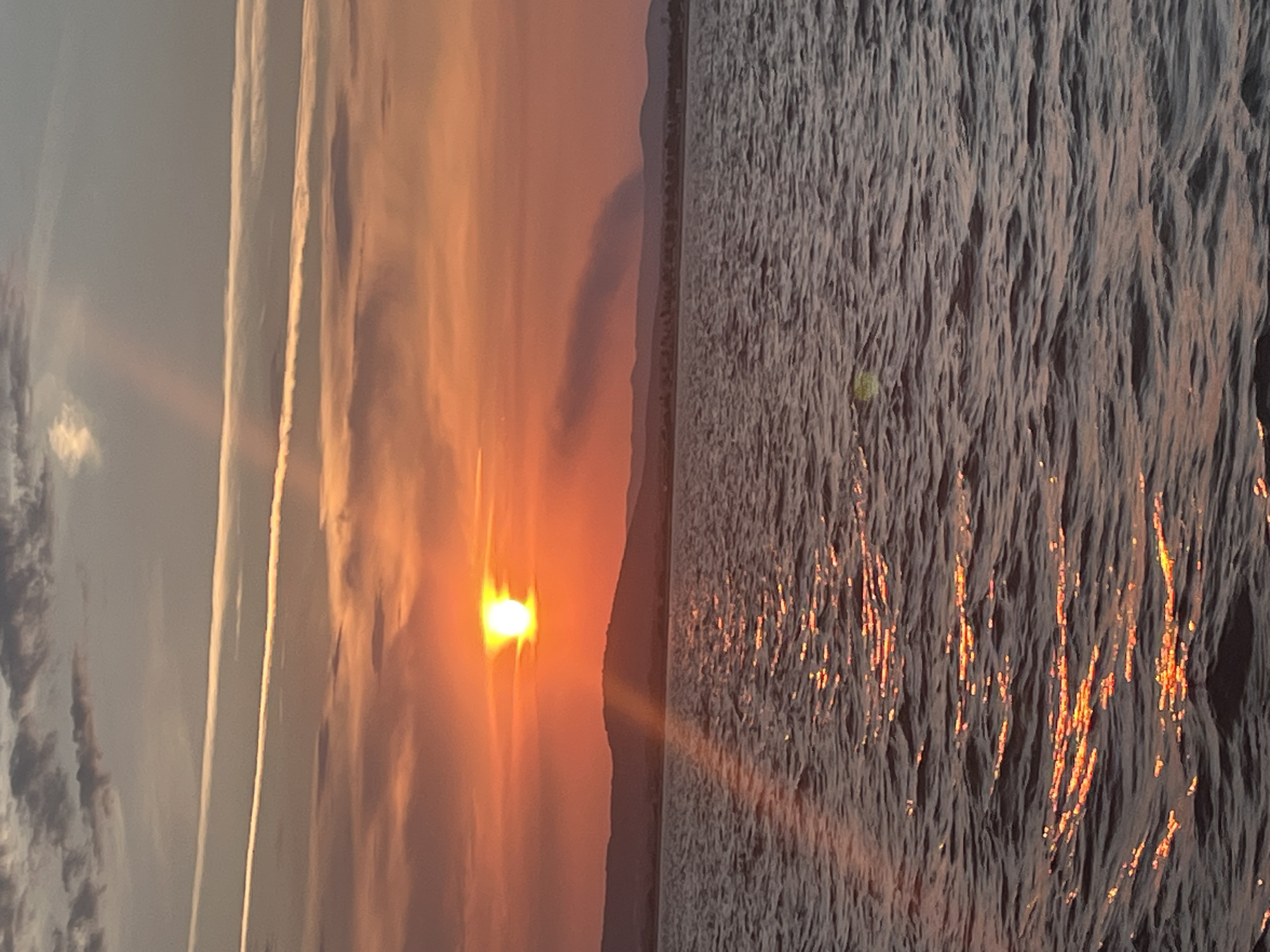 A wide shot depicts a sunset over the Mediterranean Sea near Palavas-les-Flots, France. The sun, a bright orange orb, is positioned low on the horizon, casting a warm glow across the sky and reflecting on the rippling water. The sky is a gradient of orange, pink, and gray hues, with wispy clouds streaking across it. In the distance, a silhouetted mountain range forms the backdrop, and a faint plume of smoke rises from the land. The water is dark blue with shimmering highlights from the setting sun.