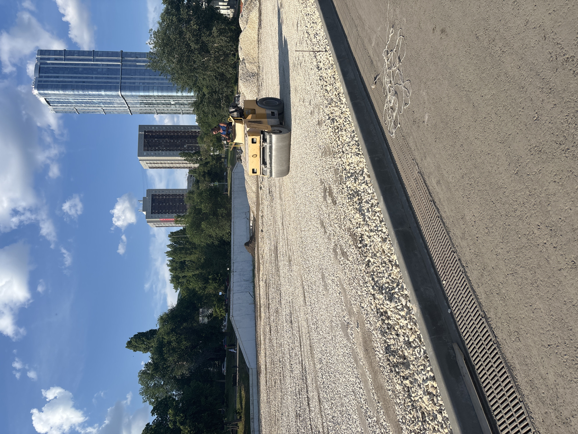 A sunny day scene captures a construction site in Saratov, Russia, with a worker operating a yellow asphalt roller on a large area covered with light-colored gravel. In the background, several tall, modern buildings, including a prominent glass-faced skyscraper, rise against a bright blue sky dotted with fluffy white clouds. Green trees line the horizon, and a concrete retaining wall is visible behind the gravel area. The foreground features a paved area with a drainage grate and a coiled white rope.