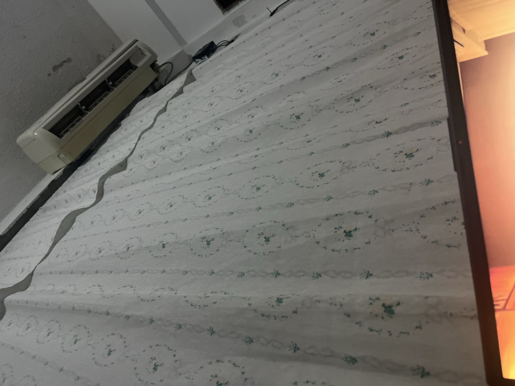 The image shows a portion of a room. A white curtain with a subtle green floral pattern covers a wall. Above the curtain, a beige air conditioning unit is mounted on the wall. The ceiling appears to be textured and shows some water stains. The lower part of the image shows the edge of a dark-framed screen, possibly a laptop or monitor, displaying a bright orange light. The scene is indoors, and no specific location in Santa Ana, Venezuela, can be identified from the visual information provided.