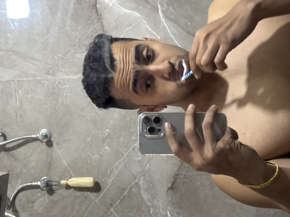 A shirtless man takes a selfie in a bathroom mirror while brushing his teeth. He holds a silver iPhone with multiple lenses in his left hand. He's wearing a gold bracelet on his right wrist. The bathroom has gray marble walls and silver fixtures. A light fixture reflects in the mirror above his head. A bar of soap and other fixtures are visible on the wall beside him.