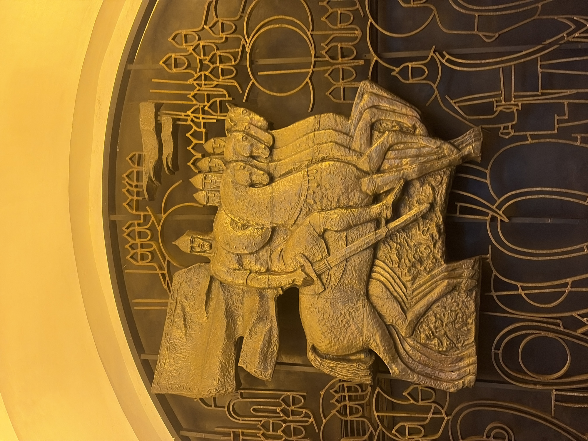 The image depicts a relief sculpture on a wall, possibly indoors, in Saint Petersburg, Russia. The sculpture is made of a gold-colored material, possibly metal or stone, and is set against a background of intricate geometric patterns. The central subject of the relief is a depiction of a mounted figure, possibly a historical or mythological warrior or ruler. The figure is riding a horse and is adorned with elaborate garments and headwear, suggesting a position of authority or status. Other figures, potentially members of a retinue, are also depicted riding horses behind the central figure. The overall style of the relief suggests a classical or historical theme, potentially related to the cultural or political history of Saint Petersburg or Russia. The scene is illuminated by artificial light.