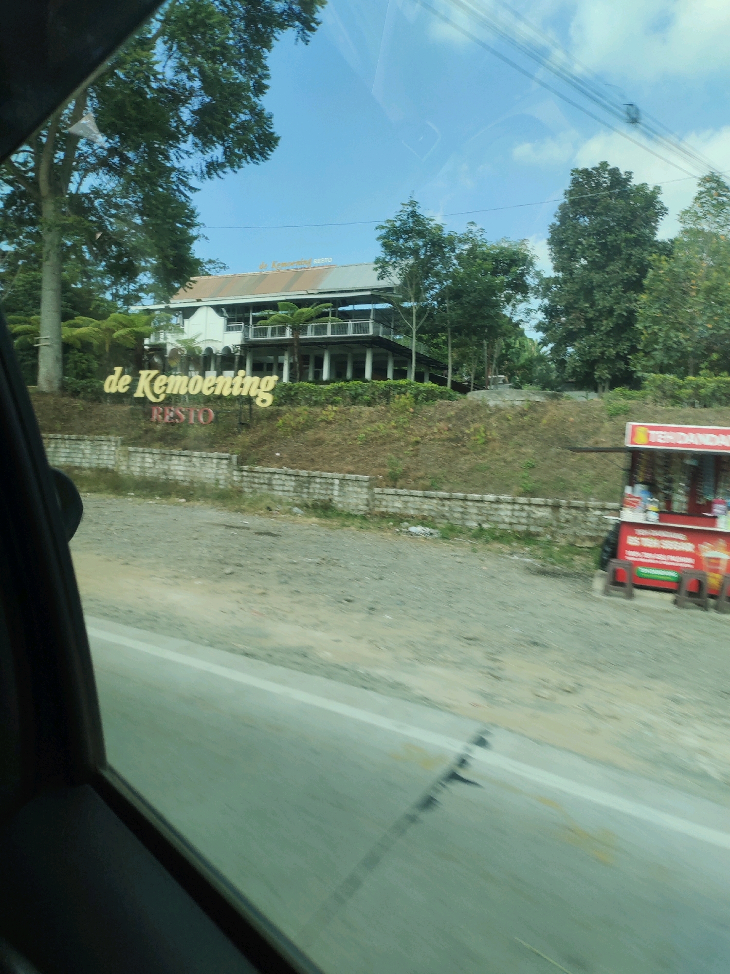 This is a photograph taken from inside a vehicle, looking out at a roadside scene in Ngargoyoso, Indonesia. In the background, a multi-story building with white columns and balconies is visible, topped with a light-colored roof. A prominent sign in large, yellow, 3D letters reads "de KEMBENING RESTO," with "RESTO" in smaller, red letters below. The building is situated on a grassy, elevated slope.
To the right of the "de KEMBENING RESTO" sign, a red and yellow roadside food stall is visible, labeled with text that appears to include "TEH DAUN DAUN" and "KOPI SEGER." Several small stools are arranged in front of the stall. The area in front of the building and the stall is a gravelly, unpaved surface. A low, grey stone wall separates the gravel area from the slope.
The foreground of the image is dominated by the interior of the vehicle, including a dark window frame and a blurred view of the road ahead, indicating motion. The sky is blue with scattered white clouds. Lush green trees surround the building and the roadside stall, suggesting a natural or semi-rural environment. The overall impression is that of a dining establishment or rest stop along a road.