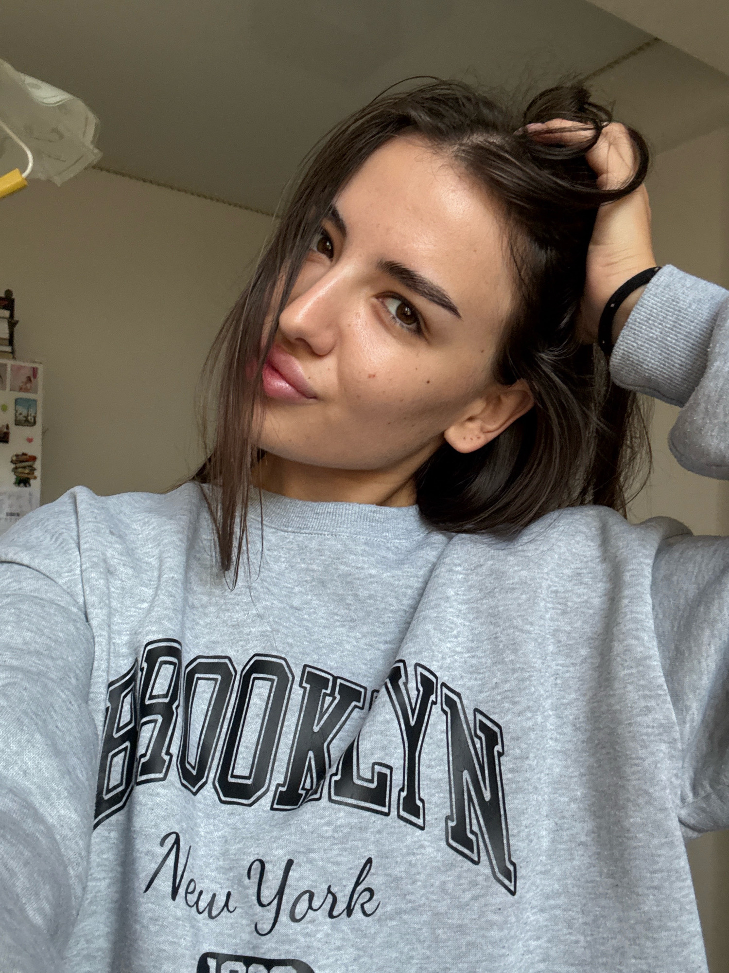 A young woman with dark hair and brown eyes is shown from the chest up, posing for a selfie. She is wearing a grey sweatshirt with "BROOKLYN New York" printed on it. Her right hand is holding a portion of her hair near the top of her head, and she is looking towards the camera with her lips slightly parted. The background is a plain, light-colored wall, with a hint of a shelf or cabinet on the left side of the image. There is no discernible location in Chișinău, Moldova.