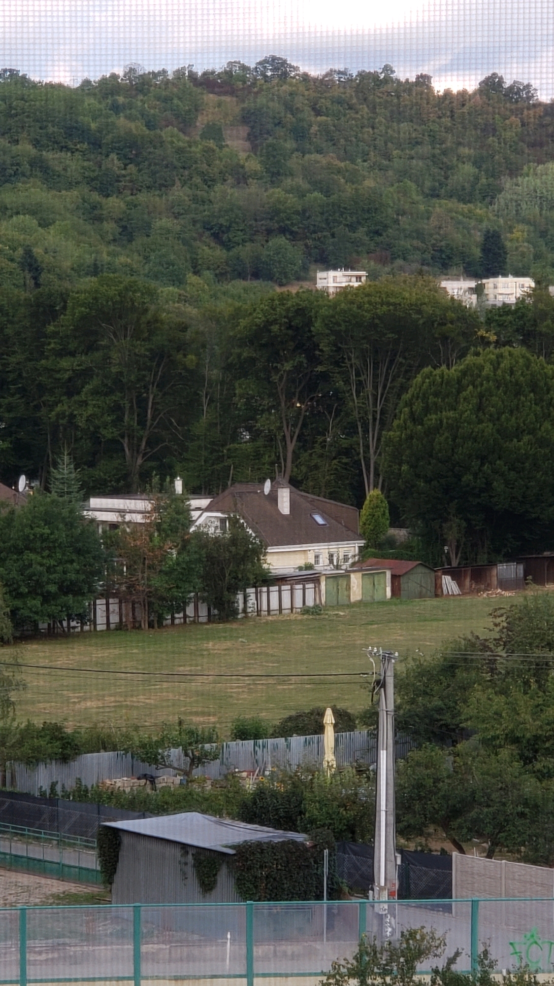 The image depicts a residential area on a hillside, with a dense forest covering the upper portion of the frame. In the mid-ground, a large, light-colored house with a brown roof is visible, surrounded by trees. Several smaller structures, possibly garages or sheds, are situated to the right of the main house. A chain-link fence runs across the foreground, partially obscuring a metallic shed and some vegetation. A utility pole with electrical wires stands in the lower right quadrant of the image. The sky is overcast. The context of Prešov, Slovakia, suggests this is a view of a neighborhood characterized by a mix of urban development and natural landscape. There are no people visible in the image.
