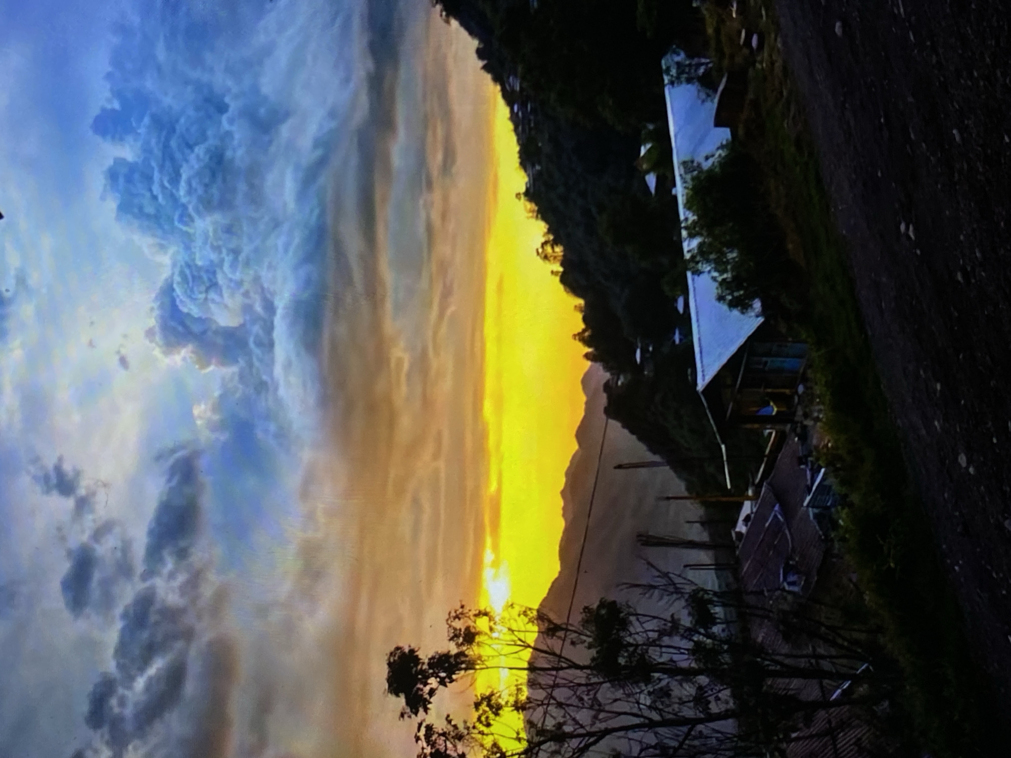 The media file displays a panoramic landscape captured during either sunrise or sunset, featuring a dramatic and colorful sky over mountains, a body of water, and scattered buildings.
The sky is a prominent feature, showcasing a clear divide in atmospheric conditions. On the left, large, textured cumulus-like clouds in shades of blue, grey, and white dominate, suggesting a cloudy area. Towards the right and center, the sky transitions into a brilliant golden-yellow and orange glow, indicating the direct light of the sun as it either rises or sets. The sun's disk is partially visible, creating an intense radiant highlight near the horizon.
A silhouette of distant mountains or hills stretches across the middle ground, sharply defined against the golden sky. Below these landforms, a calm body of water, likely a river or lake, is visible, reflecting some of the sky's ambient light.
In the foreground and midground, dark silhouettes of various trees, some with dense foliage and others with more bare branches, frame the scene. On the right side of the image, several modest buildings are situated near the water's edge, nestled among the trees. One structure stands out with a light-colored, possibly metallic, roof. Other smaller, darker structures or shacks are also present. Several vertical wooden poles are seen emerging from the water, potentially indicating an old dock or markers. A dark, unpaved road or path runs along the far right side of the scene.
The image captures a serene and beautiful natural moment, devoid of visible human activity or discernible text. The combination of the intense golden light, shadowed landscapes, and dramatic cloud formations evokes a sense of tranquility and natural grandeur, consistent with a rural or semi-rural setting such as Tinaquillo, Venezuela.