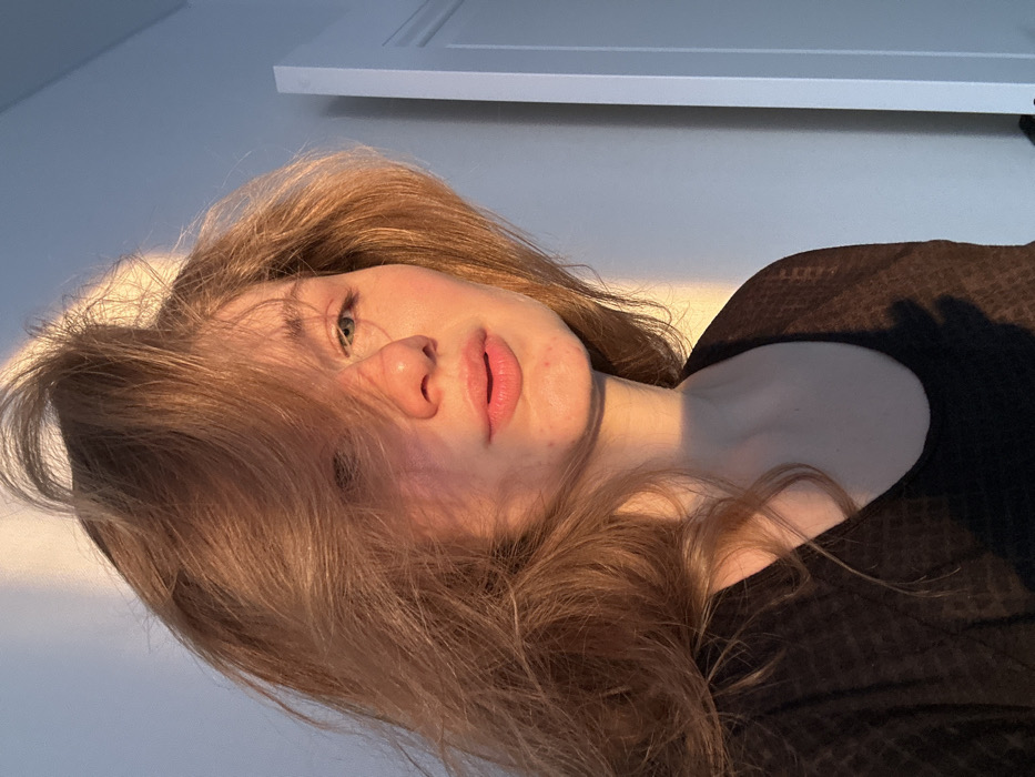 The image presents a close-up portrait of a young adult female, located indoors in Kazan, Russia. She possesses long, voluminous, light brown to auburn hair that partially covers her left eye. Her visible right eye is light-colored, and her complexion shows minor skin irregularities on the chin and right cheek. Her lips are naturally pigmented and slightly parted. She is dressed in a dark, potentially black, scoop-neck top featuring a subtle grid-like texture. Strong, warm, directional sunlight originating from the left illuminates the scene, highlighting her hair and the left side of her face and neck, while casting defined shadows, including one resembling a hand on her lower chest. The background comprises a plain, light-colored wall with a segment of a white door frame visible on the right.