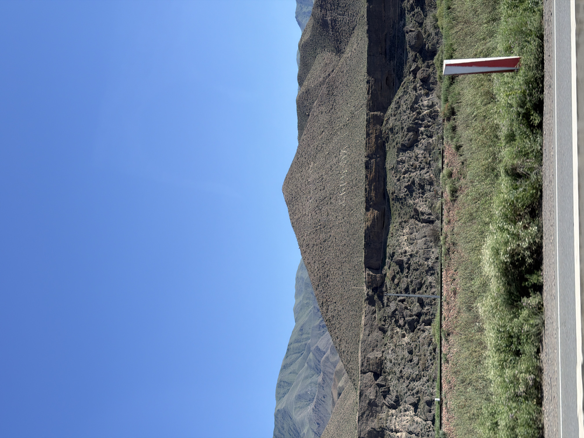 This media file depicts a vast mountainous landscape under a clear, bright blue sky, suggesting a sunny day. The scene is oriented vertically, with the sky occupying the left portion and the earth the right. The terrain is characterized by steep, rugged mountains with textured slopes covered in sparse vegetation and rocky outcrops. The foreground on the right features a paved road with white markings, bordering a strip of lush green grass and some scattered bushes. A red and white marker is visible on a pole along the edge of the road, likely a directional or safety sign. The overall impression is one of a remote, natural environment, possibly a roadside view in a mountainous region. No people or specific activities are depicted. The lighting and clear sky suggest it is daytime, likely mid-morning or afternoon. The image captures a sense of open space and natural grandeur.