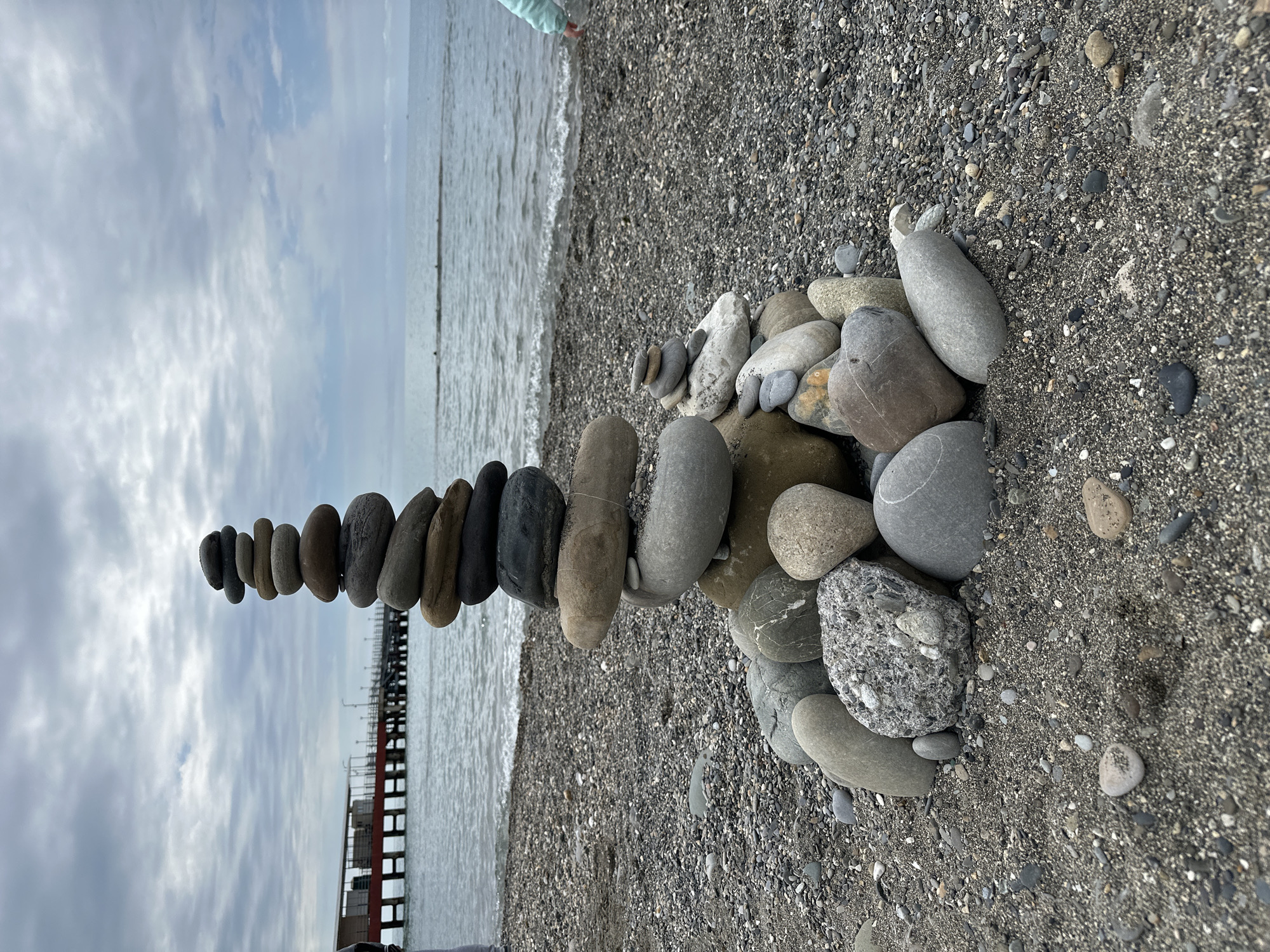 The image captures a tranquil coastal scene in Sochi, Russia, dominated by a tall, artfully balanced stack of stones on a pebbled beach. The stacked stones vary in size, shape, and color, ranging from dark grey to light brown and white, forming a slender column extending into the mid-ground. To the right of this primary cairn, several larger, smoother rocks rest on the mixed sand and gravel shoreline.
The beach itself is composed of dark, fine gravel interspersed with small pebbles, leading to the calm waters of the sea. Gentle waves are visible at the water's edge, reflecting the soft, diffused light. In the upper left corner of the original image, the lower leg of a person, wearing light-colored trousers, is partially visible near the water, suggesting human presence on the beach.
The sky is overcast with soft, scattered clouds, indicating a daytime setting with indirect sunlight. In the distance, a long pier or jetty-like structure stretches into the sea, adding a man-made element to the natural landscape. The overall mood is peaceful and reflective, showcasing both natural beauty and a moment of human interaction with the environment. No visible text is present in the image.