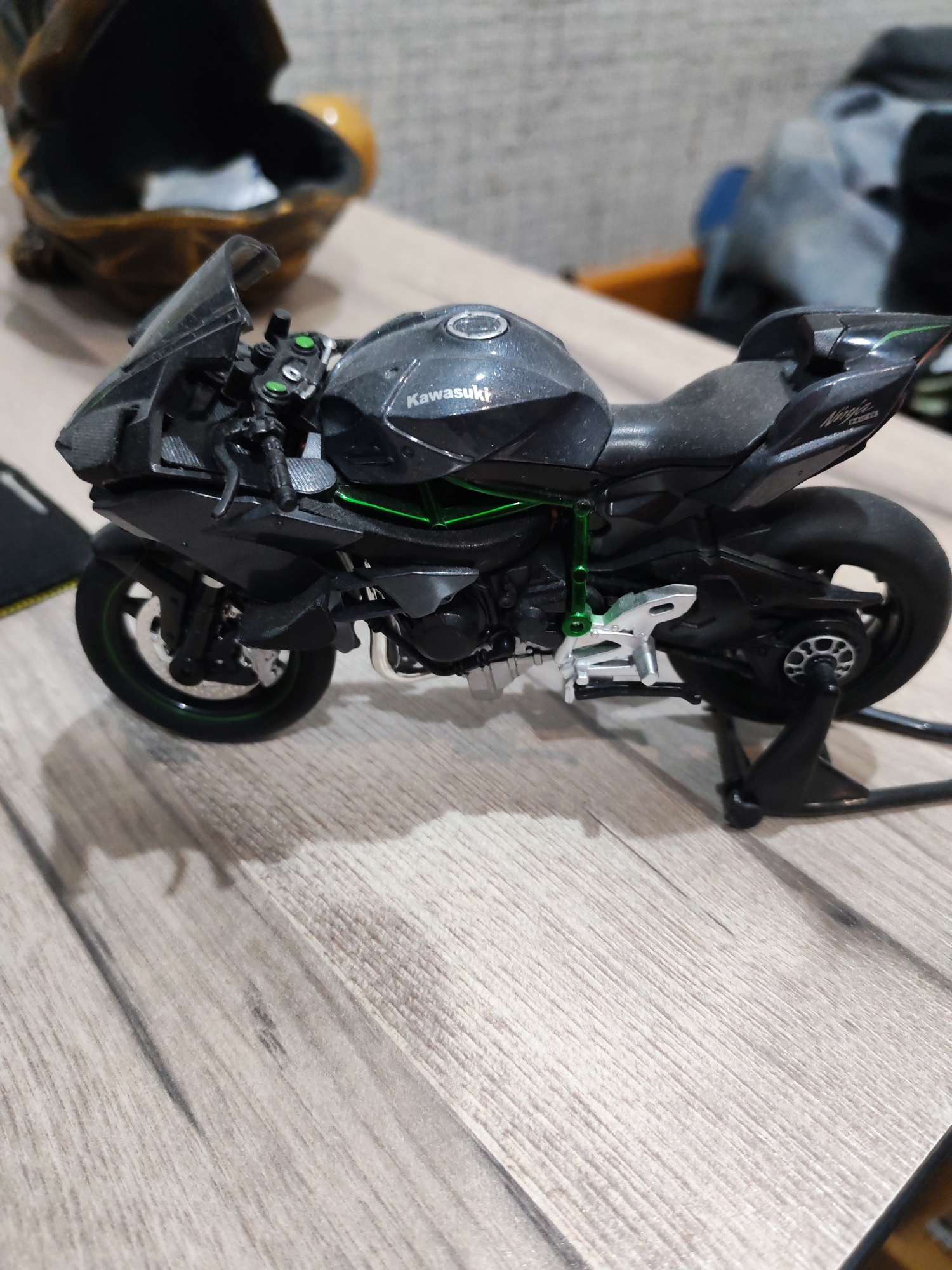 A black miniature Kawasaki motorcycle is displayed on a wooden table. The motorcycle is a detailed replica, featuring green accents on the frame and wheel rims, and the "Kawasaki" logo is visible on the fuel tank. It is supported by a black display stand. In the background, slightly out of focus, is a brown decorative object, possibly a container or sculpture, and a blurred background suggesting an indoor environment. The image does not provide any information about the location in Yalangach, Uzbekistan.
