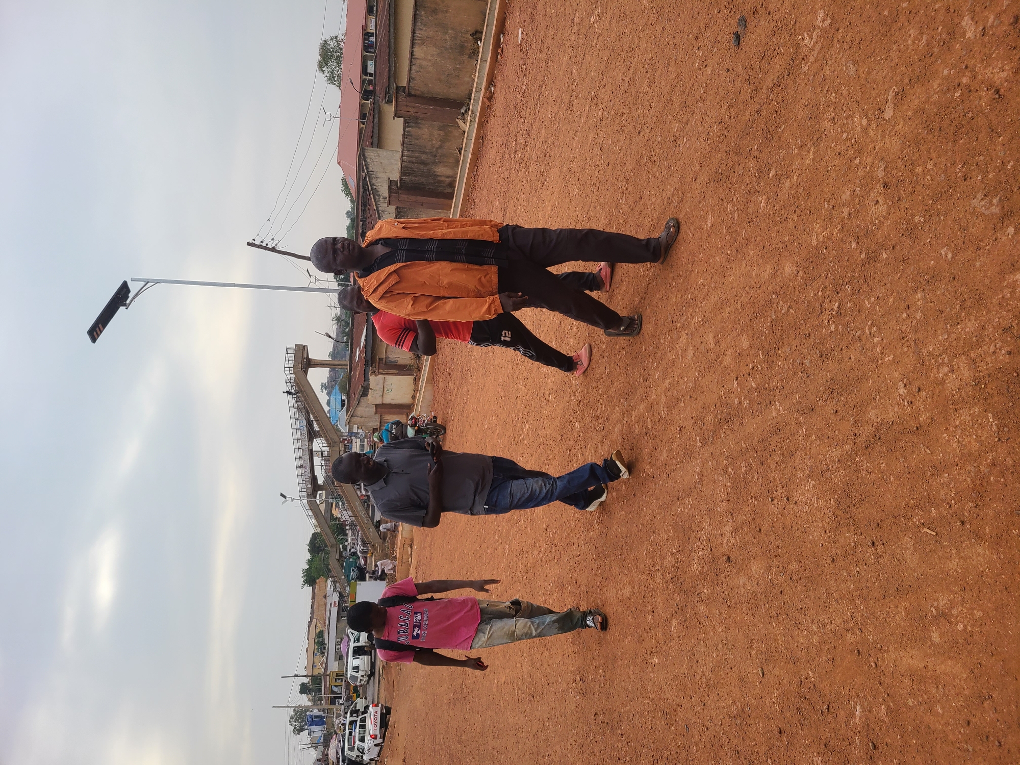 The image displays an outdoor scene in what appears to be an urban or semi-urban environment, characterized by reddish-brown laterite soil, consistent with the provided context of Jos, Nigeria. The sky is bright but overcast, suggesting daytime hours, possibly late morning or afternoon.
Four men are visible, standing or walking on the unpaved, reddish-brown ground. On the right side, a man in an orange jacket, black shirt, and black trousers walks towards the viewer. Behind him, another man in a red shirt and black trousers is also walking. Further to the left, a man in a gray polo shirt and blue jeans stands. In the far left foreground, a man in a pink t-shirt and light-colored jeans walks away from the camera; the text "JURAGAZ" and "ORIGINAL T.P.H." is partially visible on his shirt.
The background features various buildings, some with corrugated iron roofs, suggesting a mix of residential and commercial structures. A partially constructed concrete overpass or bridge is also visible. Utility poles and power lines extend across the mid-ground. On a street in the distant background, several vehicles, including a white van with "TOYOTA" visible on its rear, are present. A modern street light with a solar panel is prominent on the left side of the image. The scene depicts typical daily life in a bustling, developing area.