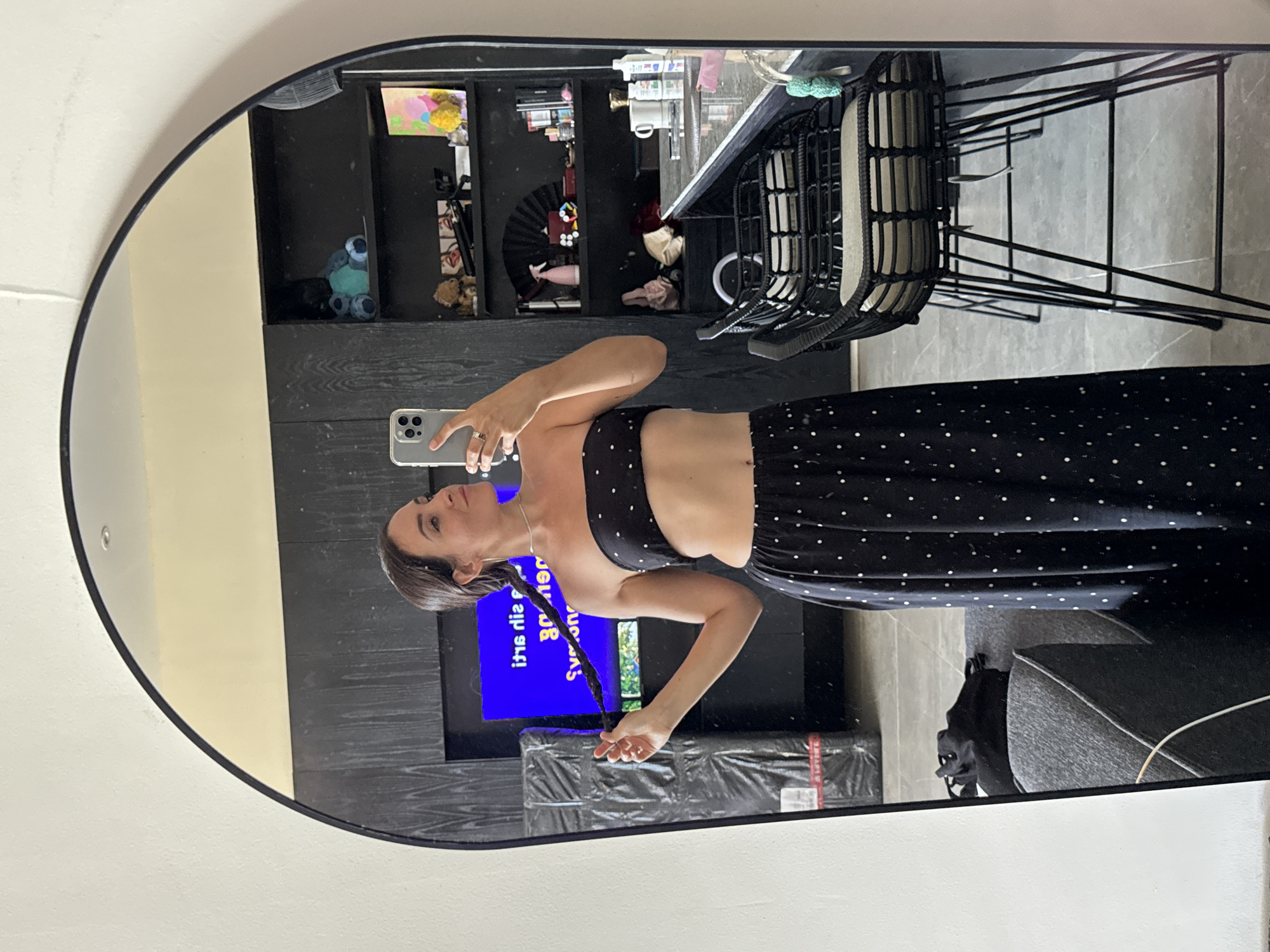 A woman stands in front of a large, arched mirror, taking a selfie. She is wearing a black strapless top and a long black skirt with white polka dots. Her dark hair is in a braid. She is holding her phone in her right hand and appears to be in a room with a television screen in the background, black shelves filled with various items, and bar stools at a counter. The reflection also shows a sofa and some wrapped items. The scene appears to be indoors, likely a home or a hotel room, and based on the reference to North Kuta, Indonesia, it is in that location.