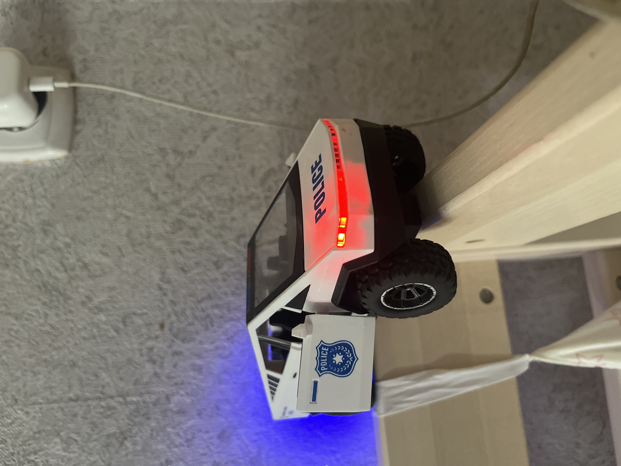 A white toy police car, modeled after a Tesla Cybertruck, is positioned on the edge of a light-colored wooden surface, possibly a bed frame. The car has its driver's side door open, revealing a black interior. A blue police emblem with the word "POLICE" is visible on the door. The car's headlights and a strip of lights on the rear are illuminated in red. Blue light emanates from the rear of the car, suggesting it may be a toy with flashing lights. The background consists of a textured grey wall. A white electrical outlet with a charging cable is mounted on the wall to the right of the car. The scene does not contain any individuals or specific geographical markers that would indicate its location within the city of Glubokoe, Kazakhstan.