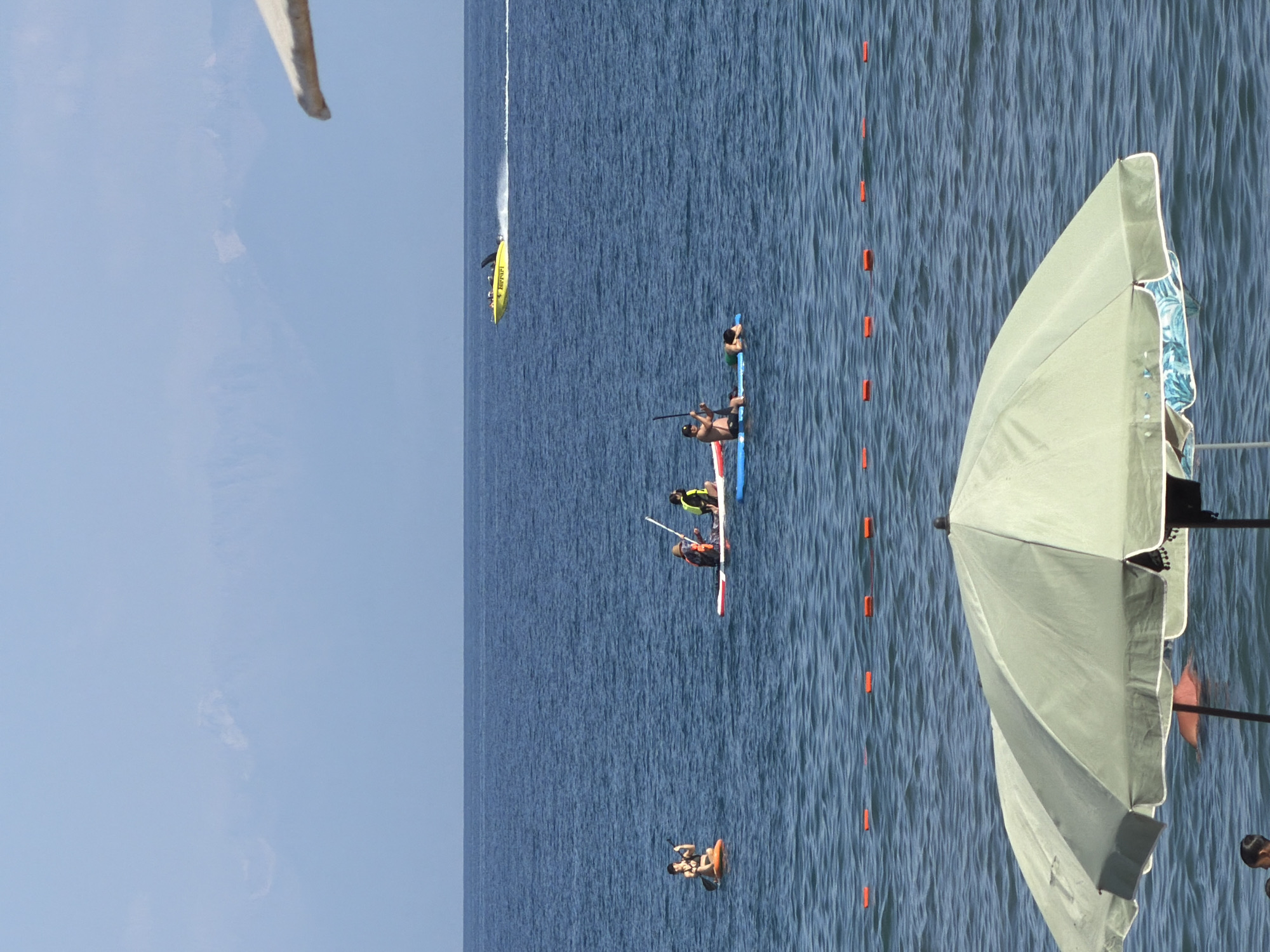 In a wide, horizontal, medium shot, the tranquil blue ocean occupies the majority of the frame under a pale sky. In the distance, hazy mountains are visible. Closer to the viewer, a yellow speedboat with "Ferrari" written on its side speeds across the water, leaving a white wake. Several people are engaged in water activities. On the left, a person is paddleboarding. Further to the right, two people are on a paddleboard, one of whom is standing. Another person is lying on a paddleboard in the water. The foreground is dominated by a large, light green beach umbrella, partially obscuring the view of the water below. A few orange buoys are spaced in a line across the water, indicating a boundary. The scene is serene and suggests a warm, sunny day at the beach.