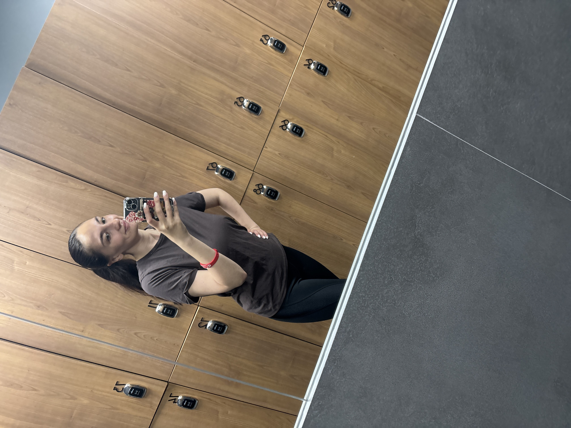 The image shows a woman taking a mirror selfie in what appears to be a locker room. The room features wooden lockers with numbered key fobs attached to each door. The floor is dark gray. The woman is wearing a dark t-shirt, black leggings, and a red bracelet. She is holding a smartphone with a patterned case. There is no indication of the specific location in Sochi, Russia.