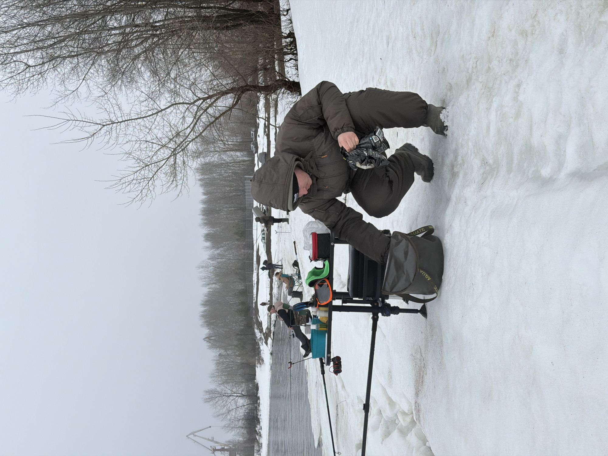 The image shows a man fishing on a snowy riverbank, likely in late winter or early spring. The setting appears to be near a river in the city of Chaikovsky, Russia, identifiable by the industrial crane visible on the opposite riverbank and the general environmental characteristics.
The man is dressed in warm, insulated clothing, including a hooded jacket and boots, presumably to protect against the cold weather. He is crouching and holding what appears to be bait or fishing tackle. Next to him is a fishing station set up on a tripod, containing various fishing accessories and equipment. Other anglers are visible in the background, also engaged in fishing along the riverbank. The environment consists of snow-covered ground, bare trees, and a river obscured by overcast skies.