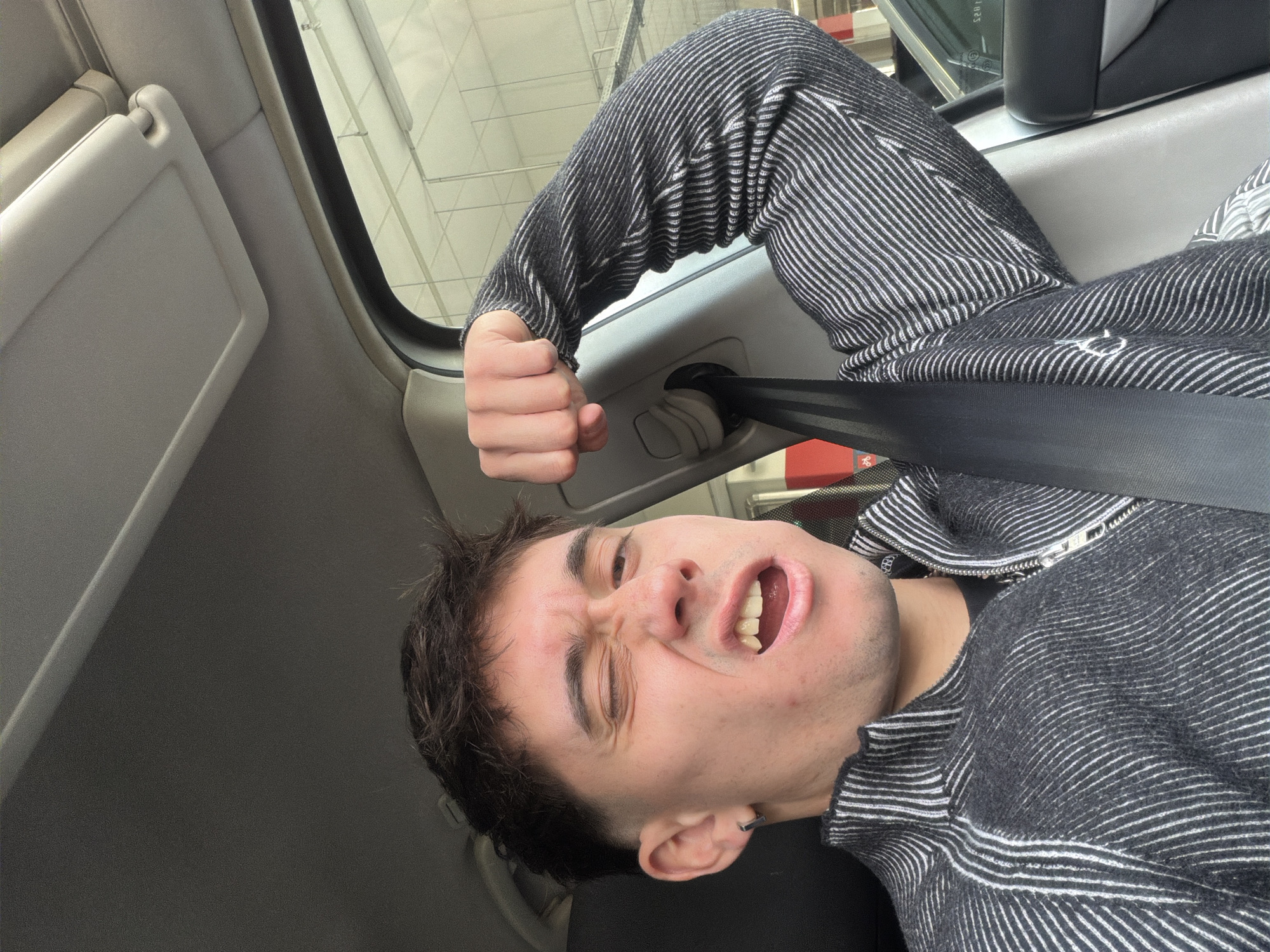 A single male individual is depicted in a close-up, chest-up shot, seated within the driver's position of a vehicle. He is wearing a dark grey or black quarter-zip long-sleeved top with fine, diagonal white stripes. A black seatbelt is fastened across his chest and left shoulder. The individual has short, dark, textured hair, a fade haircut, and a small, dark hoop earring in his left ear. He exhibits a contorted facial expression, with his left eye tightly squinted, his mouth open displaying teeth, and his nose visibly crinkled. His left arm is bent at the elbow, with the hand clenched into a fist, positioned near the vehicle's B-pillar, mimicking a flex. The vehicle's interior includes a light grey headliner and sun visor. Through the passenger window, the background suggests an indoor parking facility, characterized by fluorescent strip lighting, light-colored walls with horizontal lines, and a visible red column. The precise location within Sofia, Bulgaria, cannot be determined from the image, but the setting is consistent with an urban parking garage.