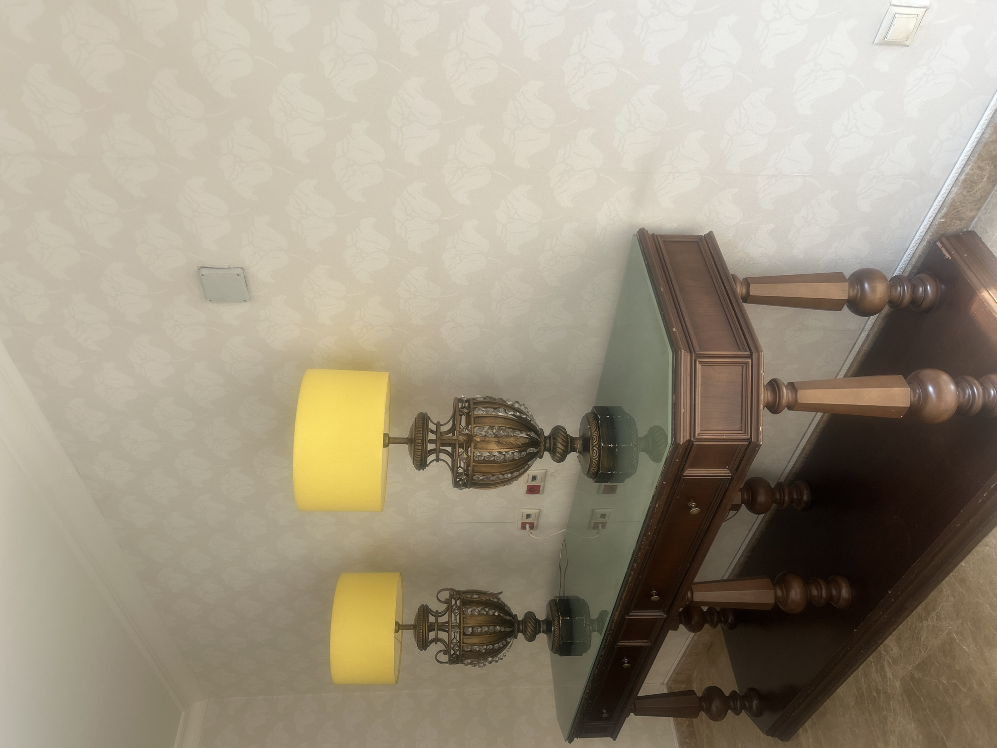 The image shows a console table with two ornate table lamps with yellow shades placed on it. The table is made of dark wood with a glass top and has several drawers. The lamps have detailed bases adorned with crystal-like elements. Behind the table, the wall is covered with light-colored wallpaper featuring a subtle floral pattern. To the right of the lamps, there are two electrical outlets and a small square electrical box mounted on the wall. The floor appears to be tiled. The scene is indoors, likely in a hallway or living space. There are no people present in the image. The lighting suggests it is daytime, with artificial light from the lamps contributing to the illumination. The overall impression is one of a well-decorated interior space, but the image itself does not contain any specific markers to pinpoint its location within Almaty, Kazakhstan.