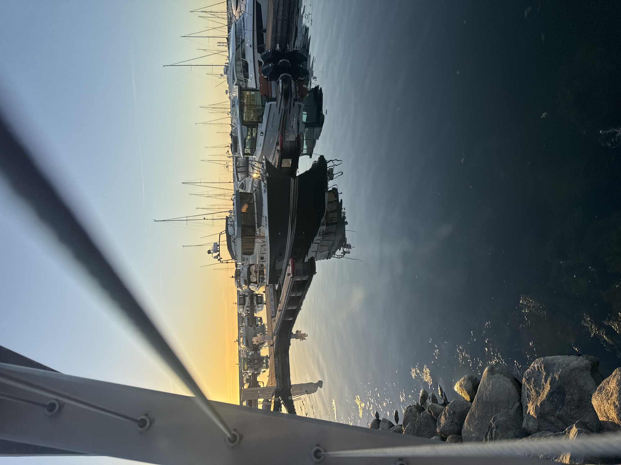 The image depicts a marina scene at either dawn or dusk in Tallinn, Estonia, specifically Noblessner Marina. The sky transitions from a bright golden-orange along the left horizon to a clear light blue in the upper right quadrant, where a faint white contrail is visible. In the immediate foreground on the left, a substantial, light grey metal support structure, possibly a pier or bridge element, angles diagonally from the bottom-left corner towards the upper-center. This structure features three visible circular bolt or cable attachment points along its vertical edge and an out-of-focus horizontal cable extending from it into the mid-frame. Below this structure, a rocky shoreline composed of large grey boulders is partially visible along the water's edge, with some rocks illuminated by the low sun.
The water surface in the marina is calm and dark, appearing deep blue-grey, with clear reflections of the sky, boats, and docks. A system of floating docks, constructed from wood or composite material, extends into the marina. A primary pier runs from the left-center towards the right, with several perpendicular finger piers.
Numerous boats are moored at the docks. In the midground, on the left, a dark-hulled motor yacht with a white cabin structure displays the registration "VKE 328" on its stern. Adjacent to it, a modern white-hulled motor yacht, identified by "SAXDOR" branding on its side, is docked with two black outboard motors visible at its stern. Both boats and their reflections are prominently featured. Further into the marina, an array of various motorboats and sailboats are docked, with the slender masts of the sailboats silhouetted against the bright horizon. No individuals are present or discernibly active within the scene.