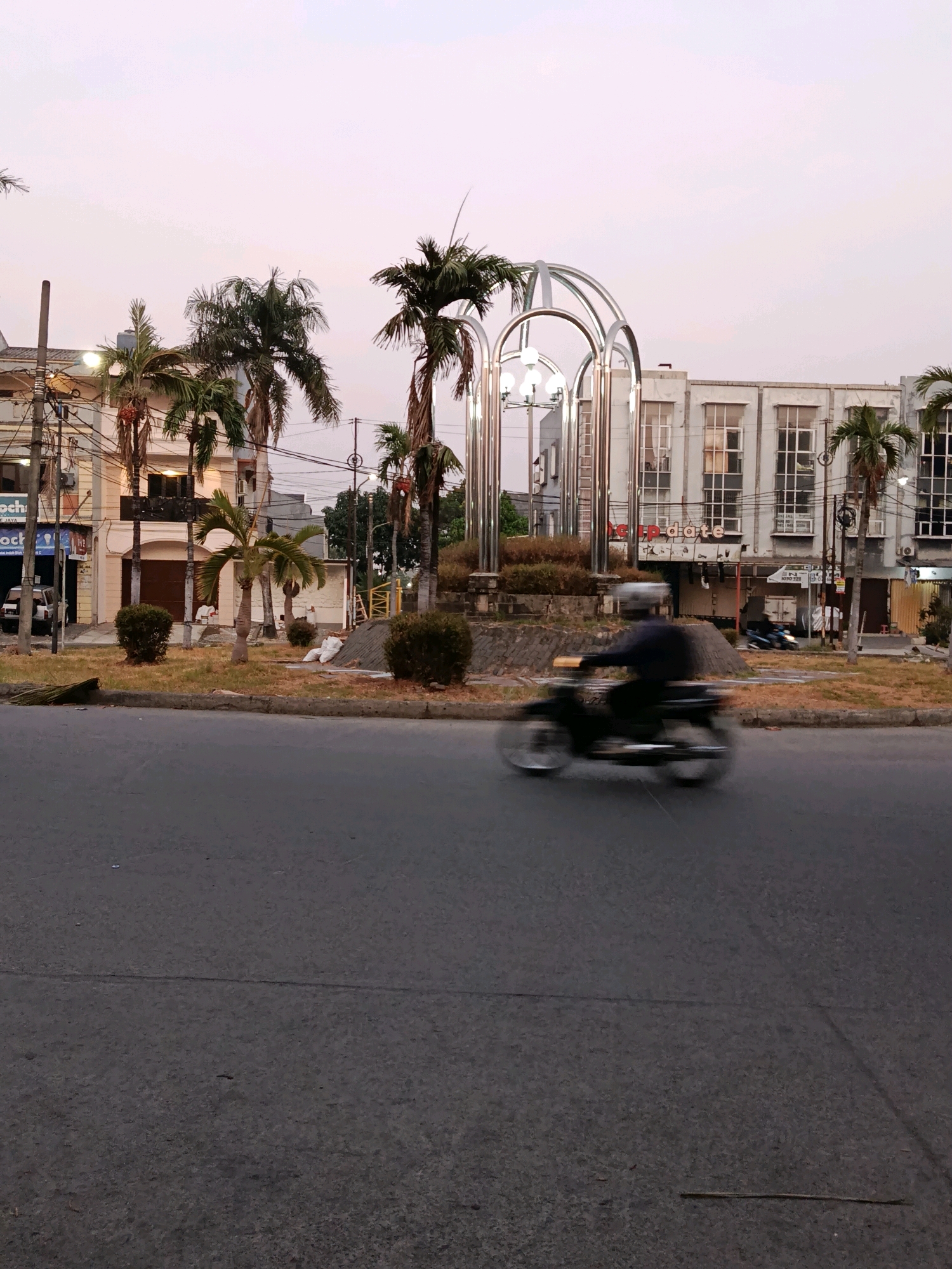 The image depicts a street scene in Bekasi, Indonesia, likely at dusk or dawn, given the soft lighting and pale sky. In the foreground, a motorcycle is captured in motion blur, indicating its movement on the asphalt road. The motorcycle has a rider wearing a helmet and a dark jacket. Behind the road, a circular median or roundabout is visible, featuring several palm trees and low-lying bushes. A modern, metallic arch structure with lights stands prominently in the center of the roundabout. Adjacent to the roundabout are buildings, including a multi-story structure with multiple windows and what appears to be shopfronts at the ground level. Some of these buildings exhibit signs with text, though not clearly legible. Electrical poles and wires are also present, typical of an urban environment. A white car is parked in front of one of the buildings on the left. The overall scene suggests a typical street in a moderately busy area of Bekasi.