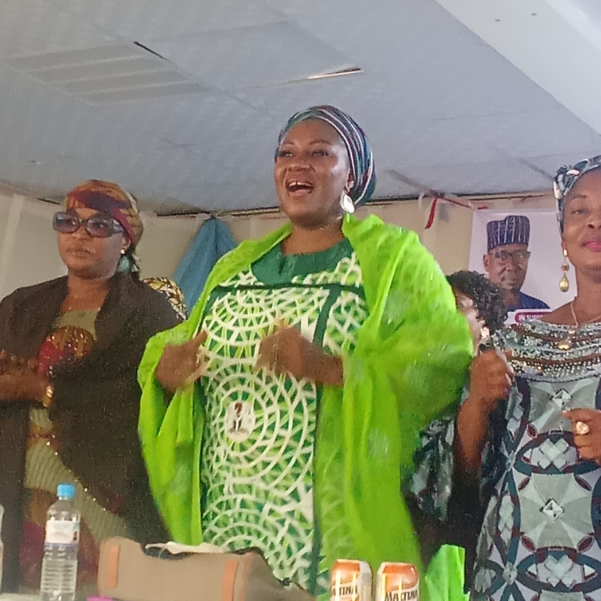 The image shows three women standing indoors, likely in a meeting room. The woman in the center wears a green and white patterned dress with a bright green shawl. She has a colorful headwrap and is gesturing with both hands. To her left, another woman wears sunglasses and a dark jacket over a patterned top, with a headwrap. To the right of the central figure, a third woman stands in a patterned dress with beaded detailing, also wearing a headwrap. In the background, partially obscured, is a poster featuring a man in a suit and traditional hat. On the table in front of the women, there are bottles of water and cans of Malta Guinness. The photo was taken in Akwanga, Nigeria.