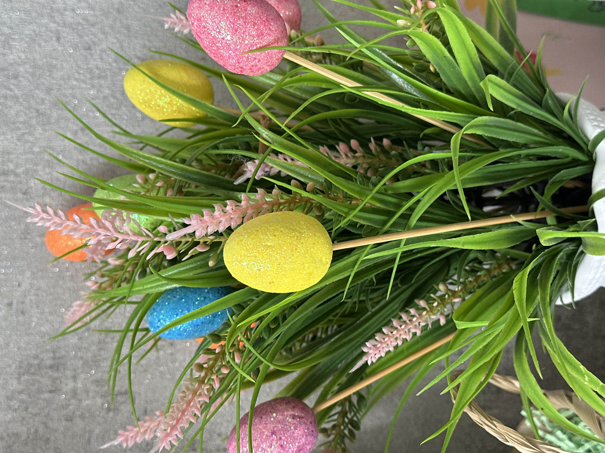 A close-up, slightly angled shot shows an Easter floral arrangement. Glitter-covered, brightly colored Easter eggs (pink, yellow, orange, blue, green) are attached to wooden sticks and interspersed among green, grassy foliage and delicate pink flowers. The arrangement appears to be in a white container inside a woven basket. The background is a gray surface with some visible sparkles. The location is not specified.