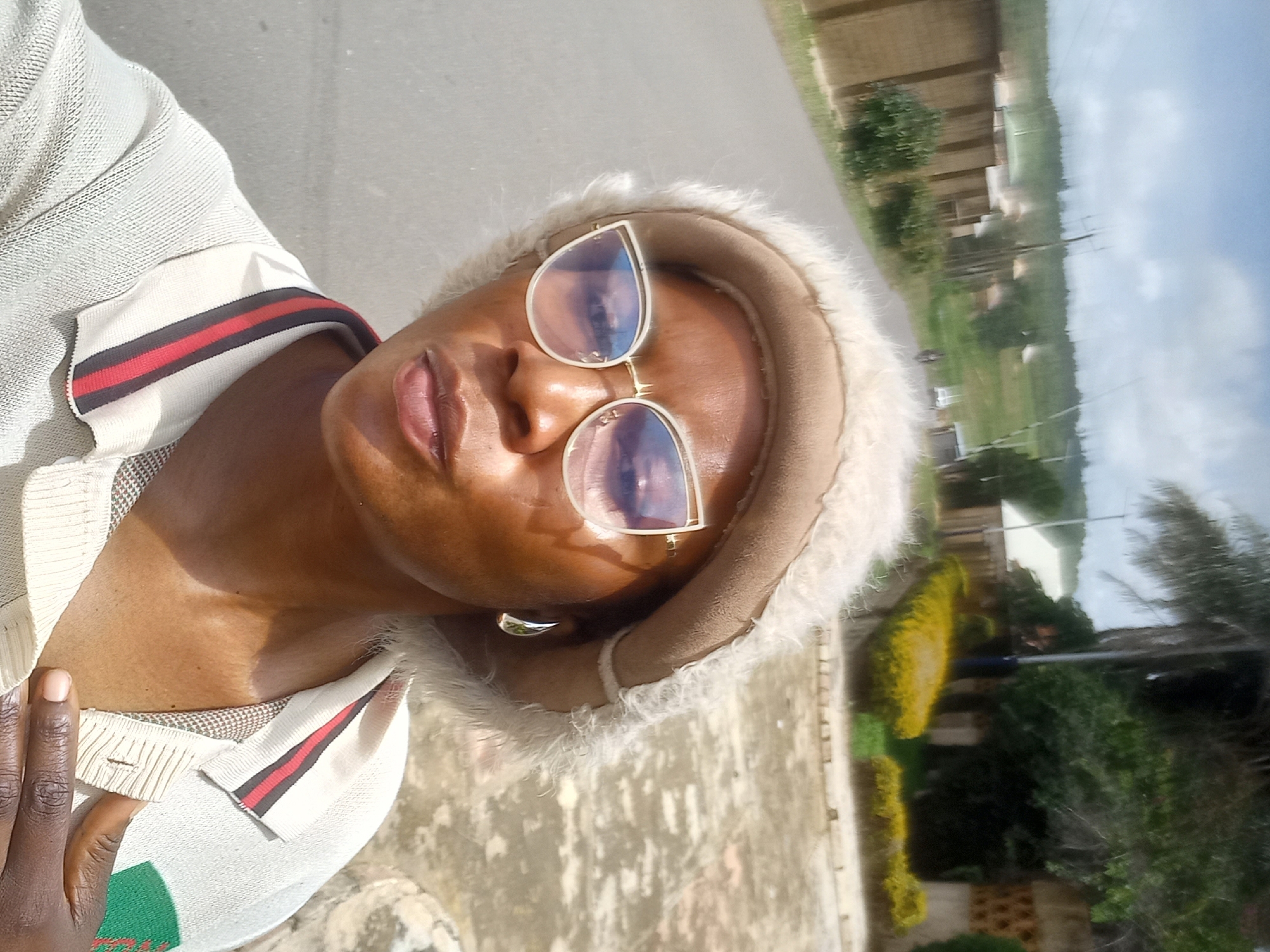 A woman wearing a fluffy tan hat and cat-eye glasses poses for a selfie outdoors. She is wearing a light-colored polo shirt with red, black, and blue stripes on the collar. In the background, a road curves uphill with houses and lush greenery on either side. The sky is partly cloudy.