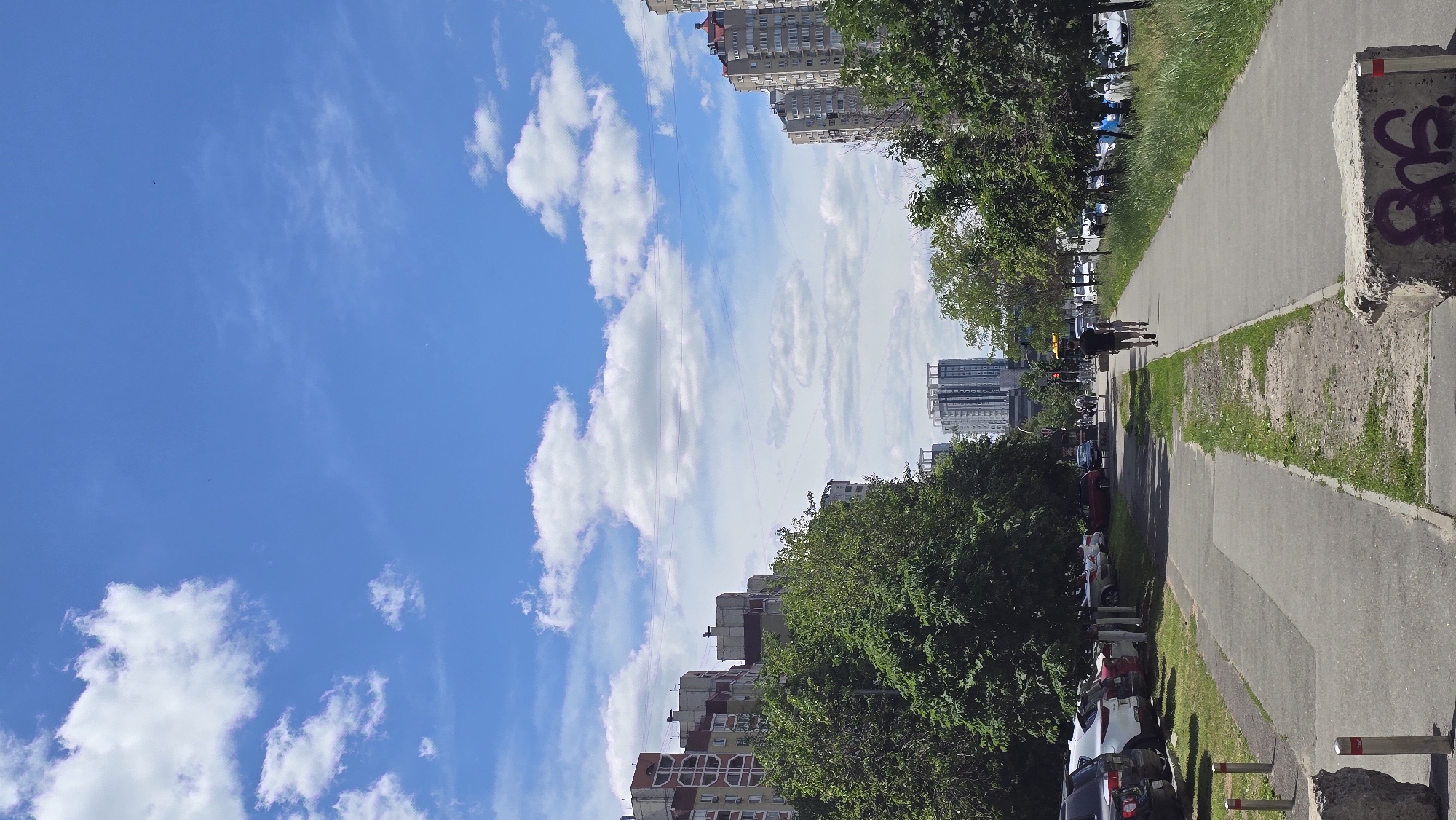 The image presents a daytime urban scene, viewed from a slightly elevated perspective. The sky, a bright blue, dominates the upper left portion of the frame, adorned with scattered white, fluffy cumulus clouds. Power lines or cables are visible stretching across the sky between buildings.
On the right side of the image, a wide, paved pedestrian path or sidewalk runs from the foreground into the distance. It is bordered by a strip of green grass on its left. Further left, a road is lined with numerous parked cars, largely obscured by lush green trees. Two individuals are walking away from the camera on the paved path in the mid-ground.
Several multi-story apartment buildings and taller skyscrapers rise in the background and along the left side of the scene, some partially hidden by the trees.
In the immediate foreground on the right, a rough, grey concrete block stands beside the path, featuring dark graffiti that appears to spell "GOS" or similar abstract characters. Further down the path on the right, two red and white bollards are visible. The scene is brightly lit by natural sunlight, indicating a clear, fair-weather day, likely late morning or afternoon.