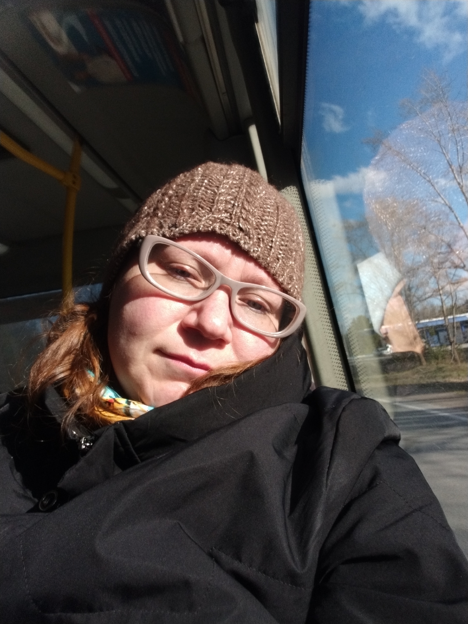 A woman wearing a brown knitted hat and glasses is sitting on a bus. She is wearing a black coat and has red hair. The bus window shows a view of a building and trees, suggesting a location in the city of Chaikovsky, Russia. The sky is blue with some clouds. A yellow handrail is visible inside the bus.