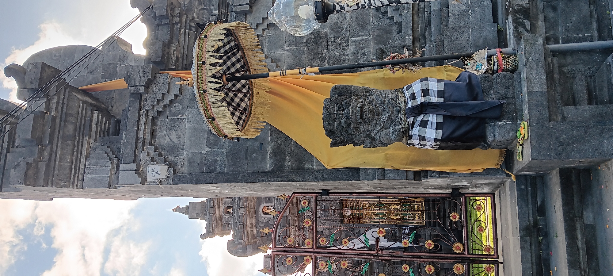 In Penebel, Indonesia, the image captures a frontal view of a stone statue situated in front of a large, textured stone wall. The statue, resembling a gargoyle or mythical creature with a prominent mouth and eyes, is adorned with a black and white checkered sarong. A yellow cloth is draped behind the statue, partially obscuring the stone wall.
To the left of the statue is an ornate, wrought-iron gate with floral motifs and yellow flower details. Beyond the gate, a glimpse of green grass and a tiered, stone temple structure with a pointed roof can be seen. In the background, the sky is partly cloudy.
Above and to the right of the statue, a decorative umbrella with a black and white checkered pattern and yellow fringe is held aloft by a black pole. A glass lamp is visible on a pole to the far right of the image. At the base of the statue, on a small stone ledge, are two small offering baskets containing green leaves and what appear to be red flowers. Steps are visible leading up to the statue and the gate, made of dark stone. The overall scene suggests a cultural or religious site.