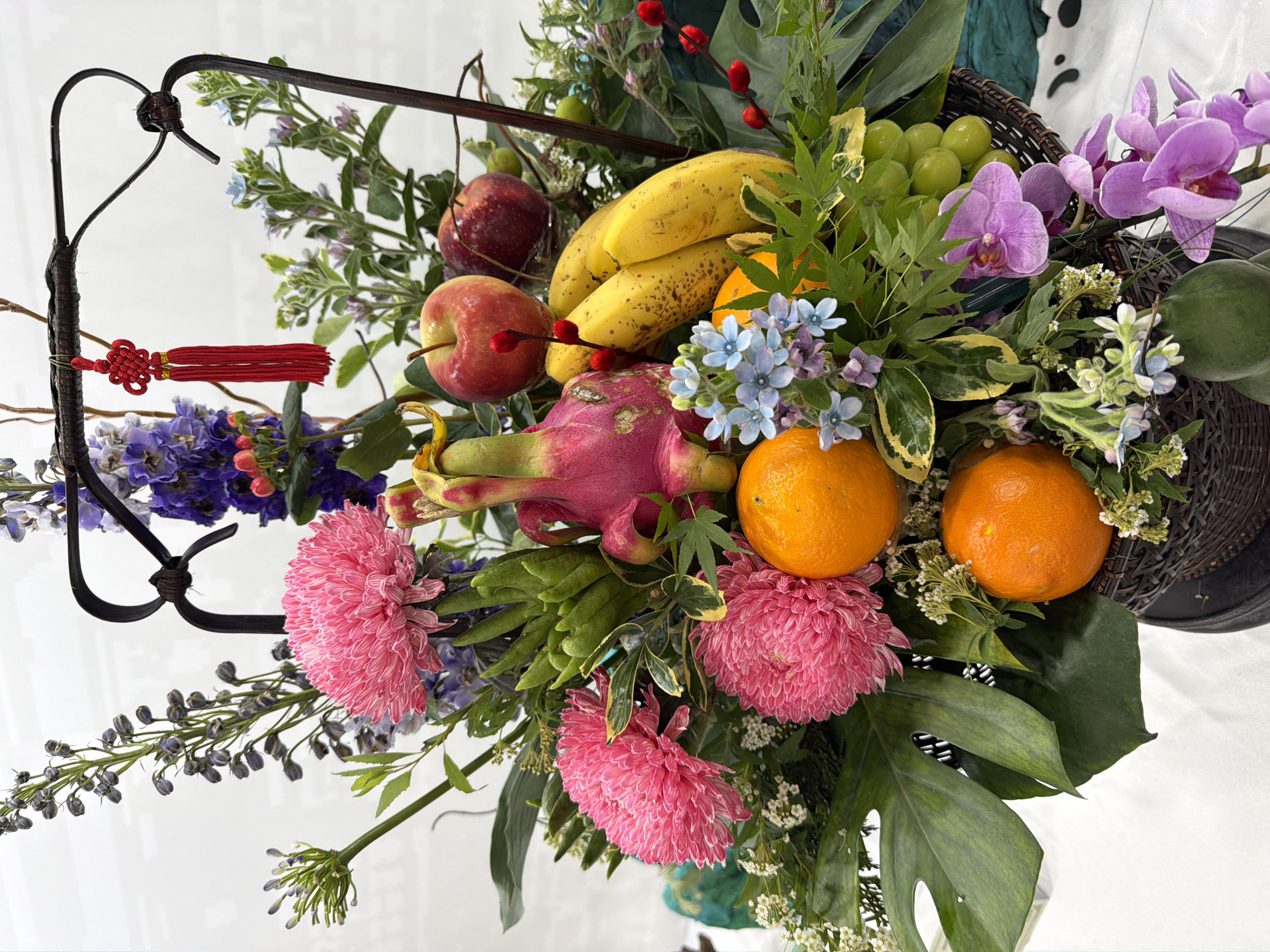 A lush floral and fruit arrangement is displayed indoors. The arrangement includes pink chrysanthemums, purple orchids, blue forget-me-nots, green leaves (possibly Monstera), red apples, yellow bananas, oranges, green grapes, and a dragon fruit. The arrangement is contained within a dark woven basket with an attached ornate metal framework. A red Chinese tassel hangs from the metal framework. The setting is a bright, neutral background, likely a tabletop or display surface. There is not enough information to place the image in Haizhu District, China.