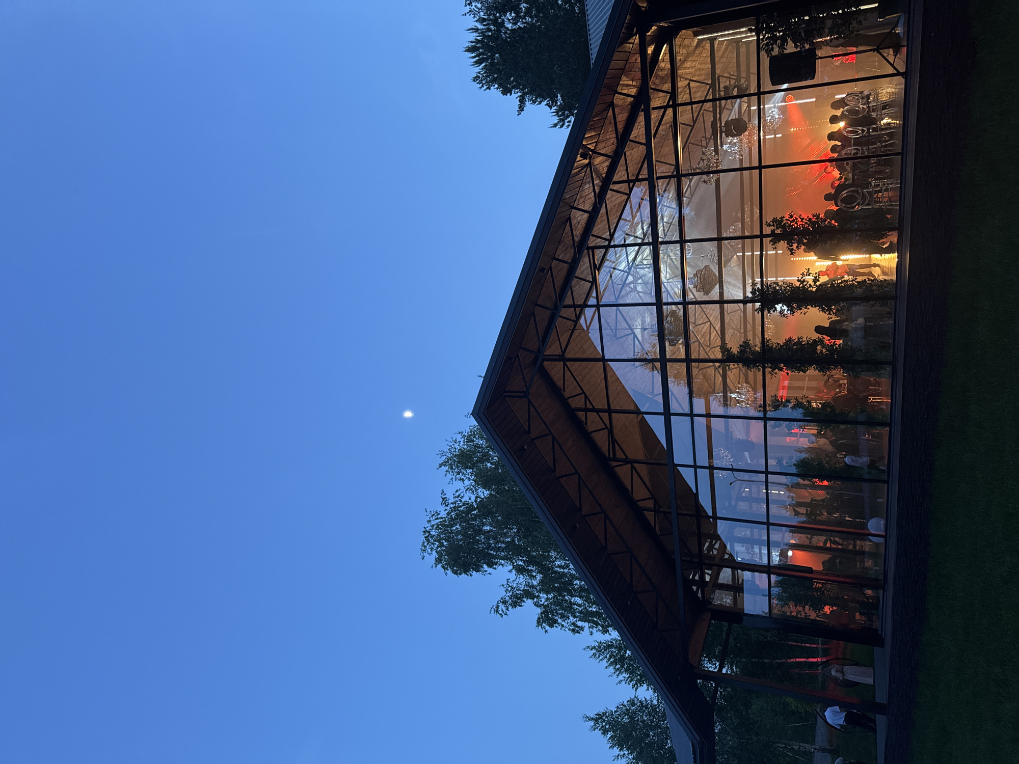 The image depicts a modern glass-walled building at dusk in Эстачи, Russia. The sky is a deep blue with a faint moon visible. The building has a gabled roof structure, and its interior is lit with warm, orange and red lighting. Inside, rows of chairs and tables are set up, suggesting an event or gathering. Dark silhouettes of people are visible through the glass. Trees are visible around the building.