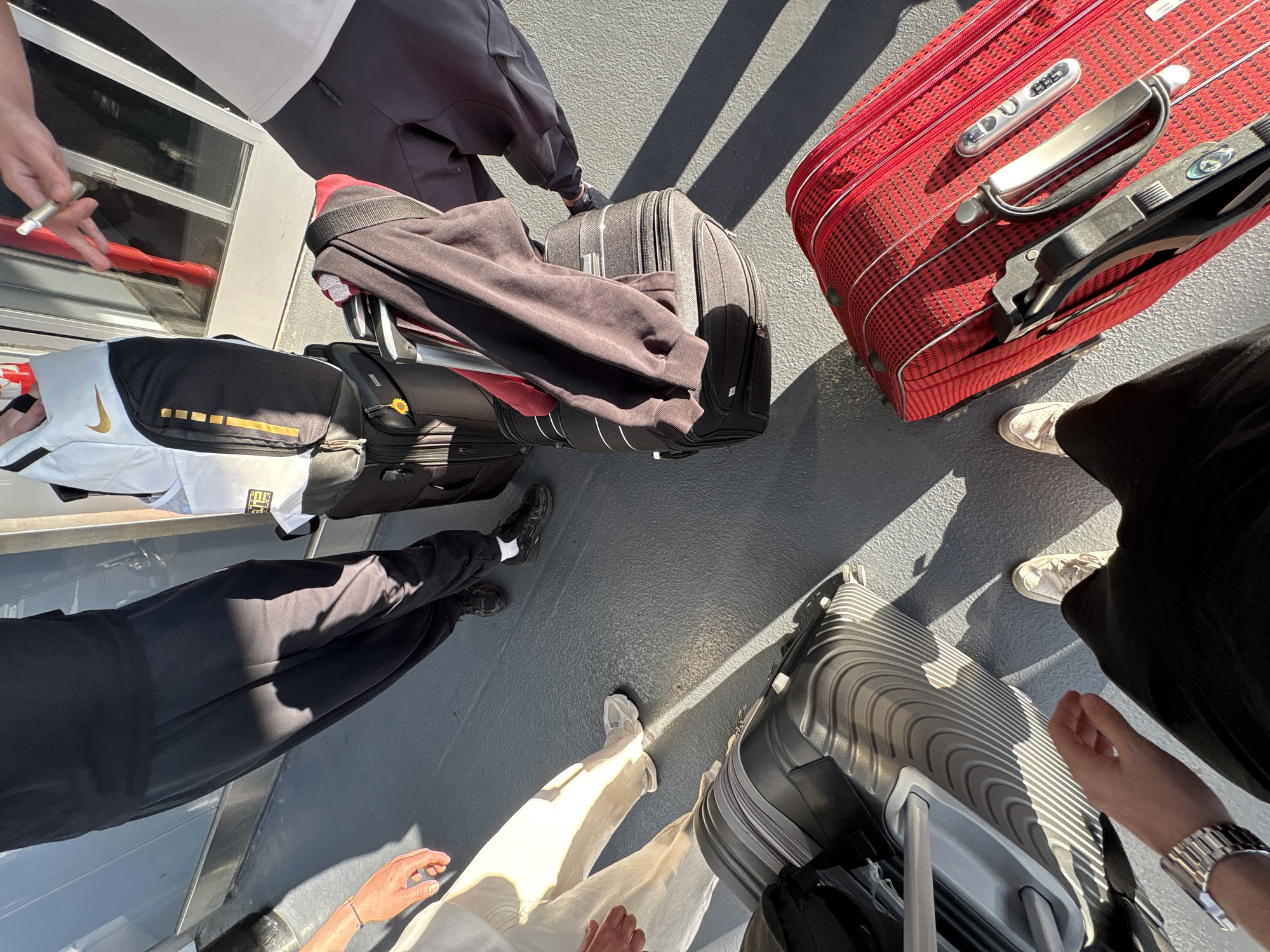The image shows a top-down view of several people and their luggage on what appears to be a ferry or boat deck. There are four pieces of luggage visible: a large red suitcase with a telescopic handle, a black rolling suitcase, a black bag with white and gold detailing on top of a grey suitcase, and a silver rolling suitcase. Four people are partially visible, wearing varied clothing styles, including black pants, light-colored pants, and a dark top. One person has a silver watch on their wrist. The setting seems to be outdoors, indicated by the natural lighting and shadows. The location in College Park, United States of America, cannot be confirmed from the visual information provided.