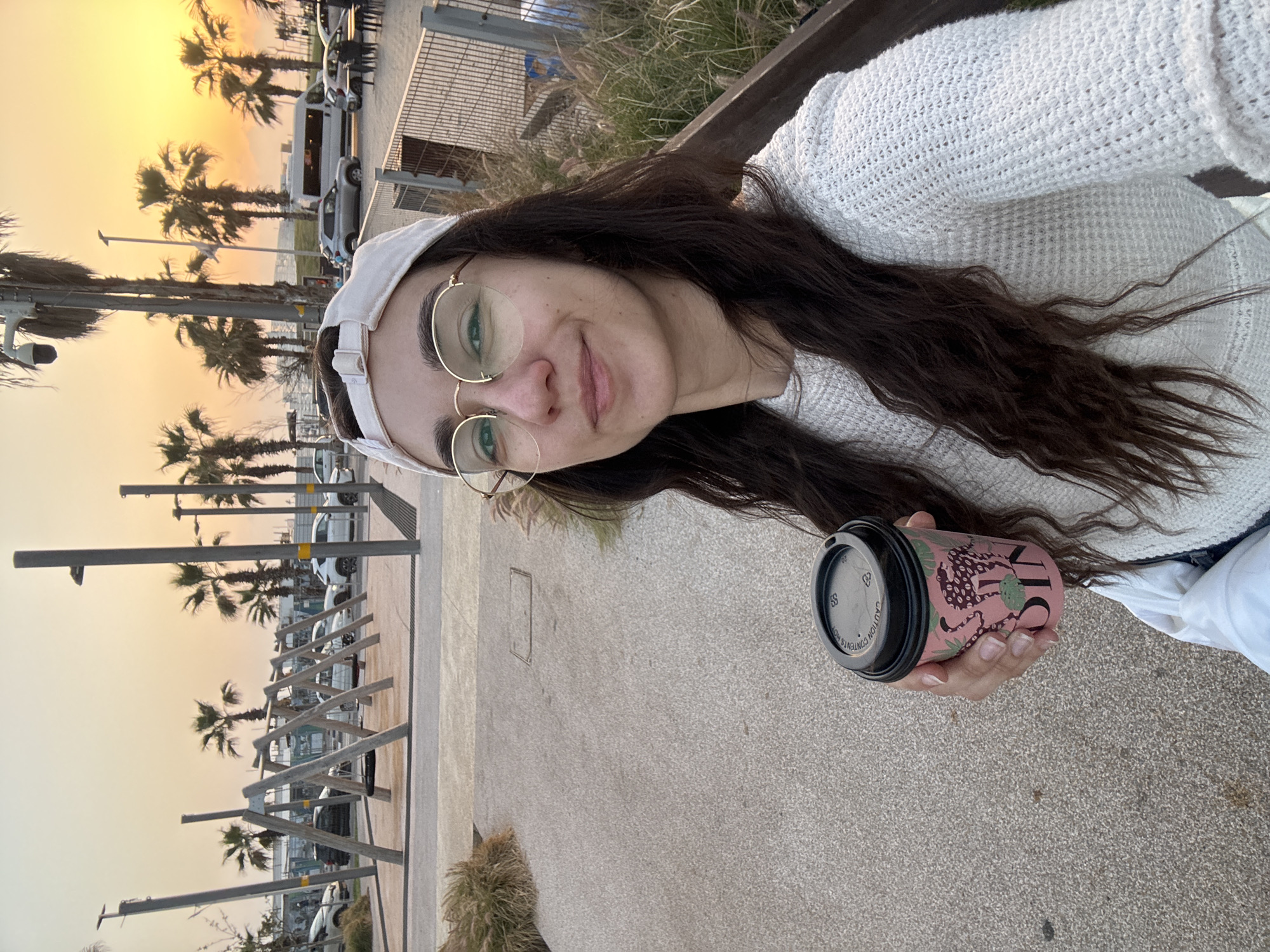 A woman is taking a selfie outdoors at sunset, possibly in Bat Yam, Israel. The woman has long brown hair, wears a white sweater, round glasses, and a white baseball cap. She's holding a pink paper cup with a black lid, likely a coffee cup. Behind her, palm trees and streetlights are visible against a bright orange sunset. A row of parked cars are also in the background. The ground is paved. There are metallic structures nearby and some sparse vegetation.