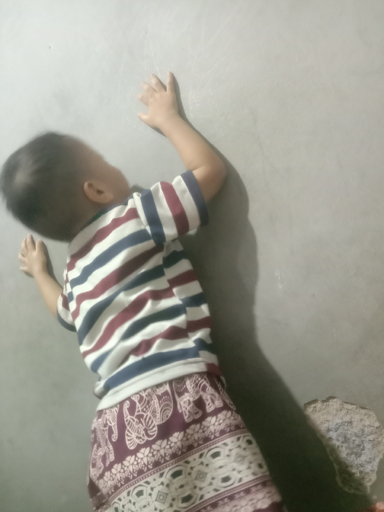A young child, viewed from behind, leans against a grey wall. The child wears a horizontally striped t-shirt with bands of white, dark blue, and maroon. Below the t-shirt, the child's lower body is covered by a maroon garment patterned with white elephants and floral designs. The child's right hand is extended, touching the wall. A shadow of the child is cast on the wall to the left. To the right of the child, a rough, light-colored stone is visible near the base of the wall. The setting appears to be an interior space with a concrete wall.