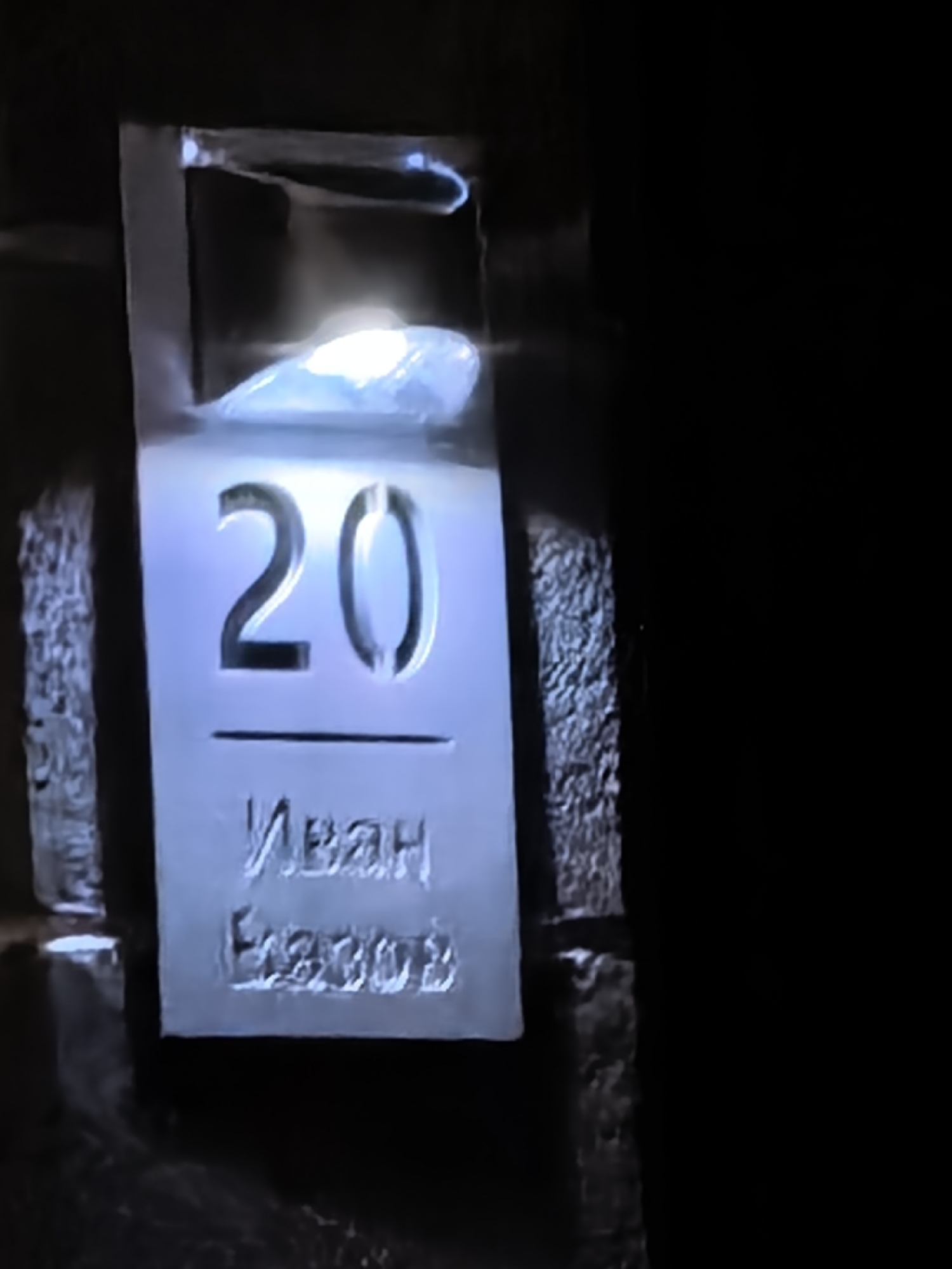 A sign on a wall reads "20" above a horizontal line, followed by the Cyrillic script "Иван Елзов." The sign is illuminated, and the immediate surroundings are dark. The location is Unknown, Unknown.