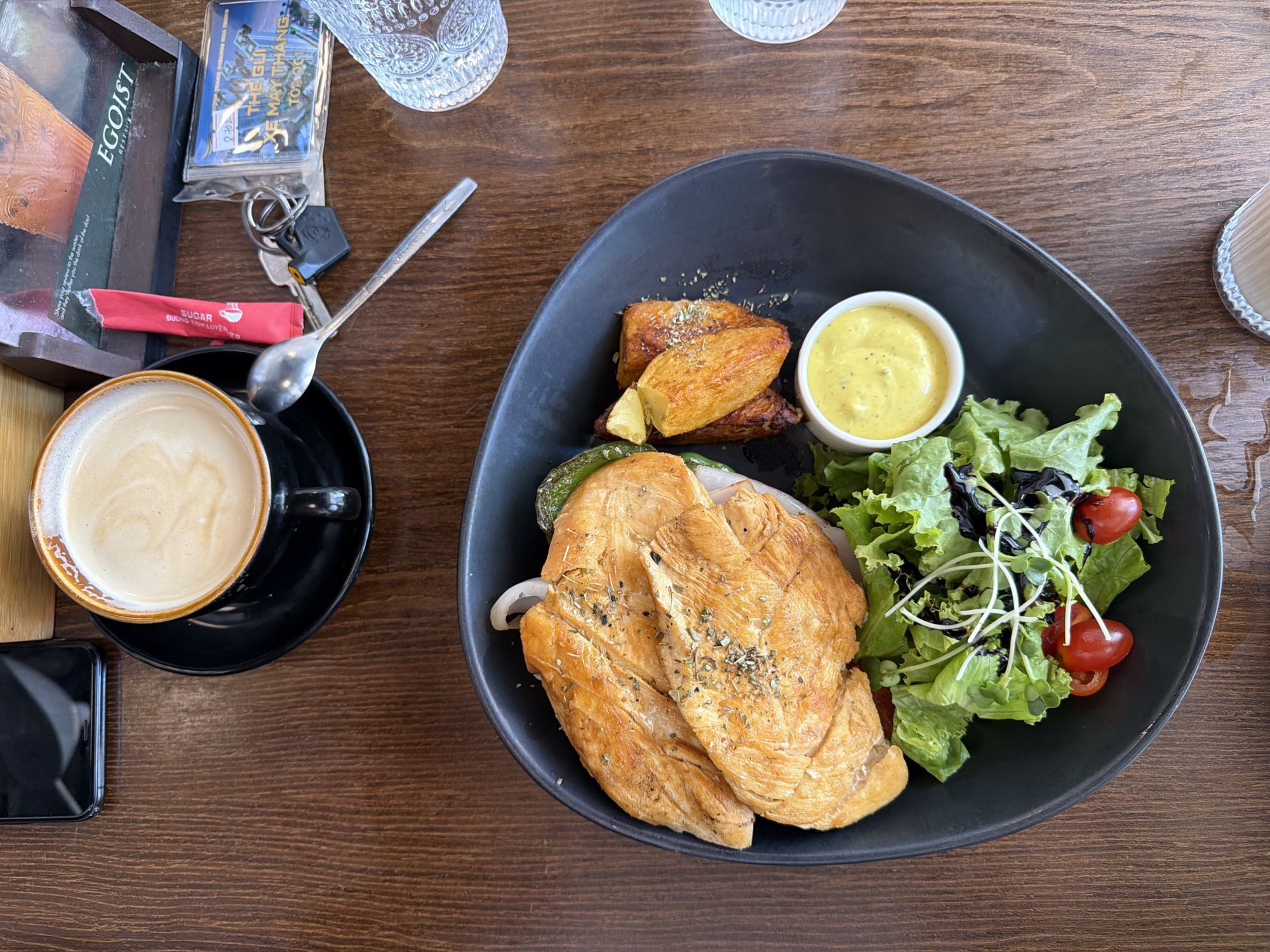 An overhead shot captures a meal and coffee on a wooden table, likely at a cafe or restaurant in Nha Trang, Vietnam. The main dish, served in a dark, oval bowl, consists of grilled or pan-fried chicken breasts topped with herbs, accompanied by roasted potato wedges, a side of fresh green salad with cherry tomatoes and sprouts drizzled with balsamic glaze, and a small cup of yellow sauce, possibly a mustard or aioli.
To the left of the meal, a black cup of coffee with a light froth sits on a black saucer. Next to the coffee, a dark-colored smartphone is visible. In the upper left corner, various personal items are arranged on a wooden tray with a partial inscription. These include a red sugar packet, a set of keys with a black fob, and a clear plastic bag containing what appears to be a card. A spoon rests near the sugar packet and coffee cup.
Visible text includes "EGOIST" on the wooden tray, "SUGAR" and "ĐƯỜNG TINH LUYỆN" (Vietnamese for "refined sugar") on the red packet, and "THE GUY," "XE MÁY THÁNG" (Vietnamese, possibly relating to a monthly motorbike pass), and "TOANQG" on the item in the plastic bag, along with the number "2702." Partial English text "Show y fer you the drink of the day!" is also visible on the tray. Two decorative clear glass tumblers are partially visible on the table. The scene suggests a relaxed daytime meal.