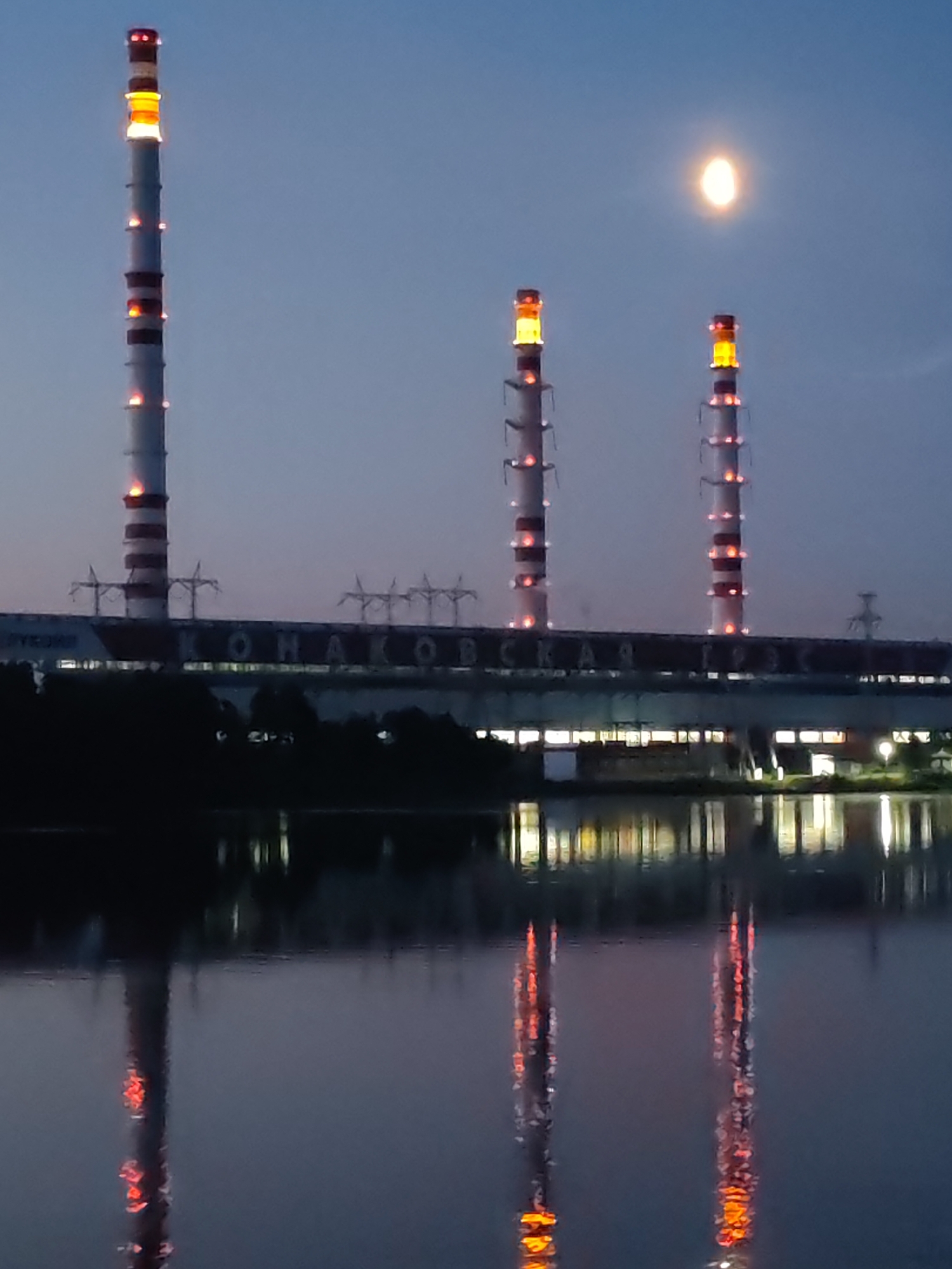 The image depicts a night scene at a power plant. Three tall smokestacks, illuminated with red and orange lights, dominate the left and right sides of the frame. The central smokestack is obscured by a building. The building, which appears to be part of the power plant, has a long, horizontal sign with Cyrillic lettering. The sky is a deep blue, with a bright, oval-shaped object, possibly the moon, positioned above the central smokestacks. In the foreground, a body of water reflects the lights of the smokestacks and the building. Trees are visible along the edge of the water, silhouetted against the night sky. The overall impression is one of industrial infrastructure set against a natural landscape at dusk or night.