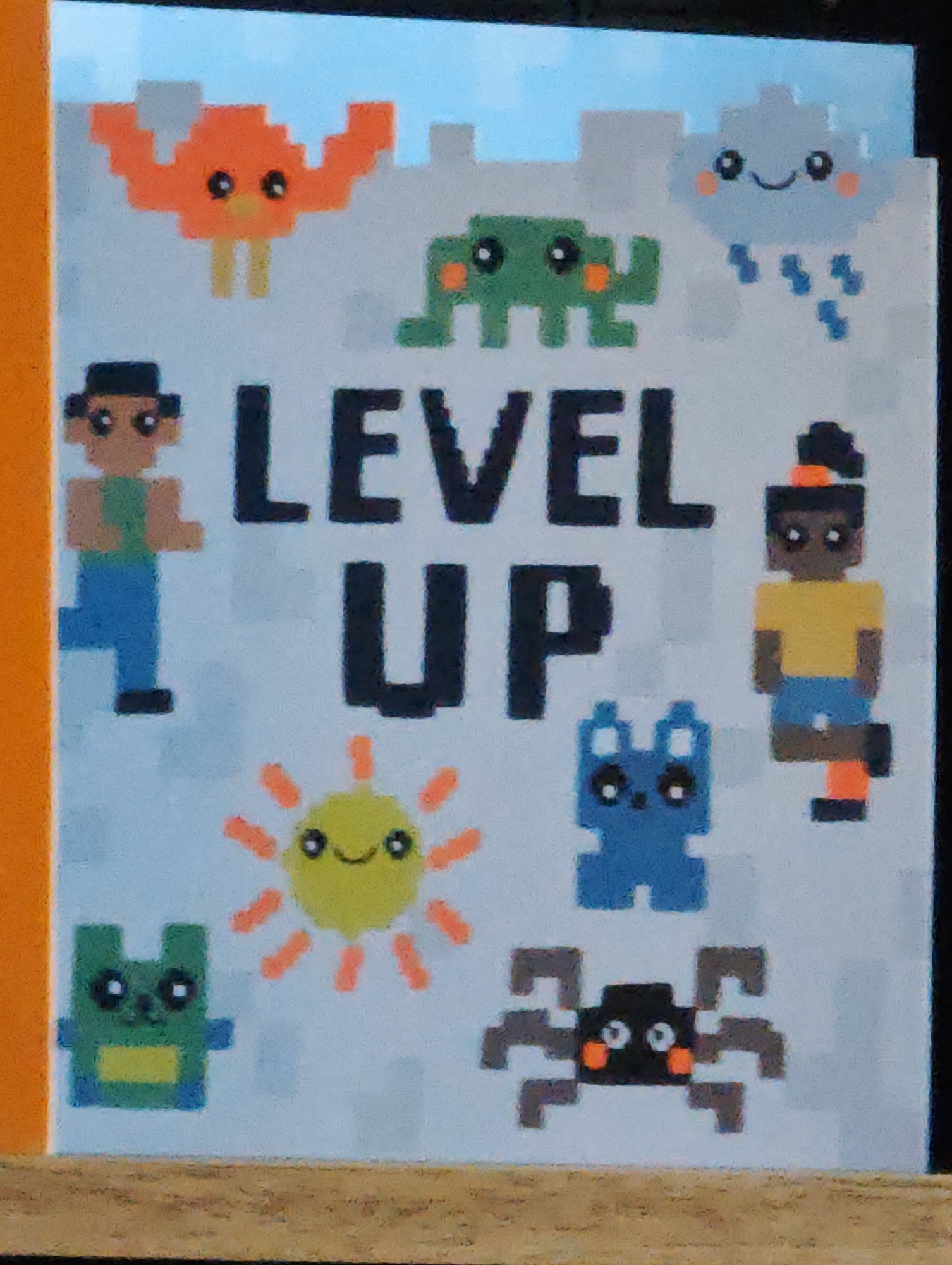 The image shows a close-up view of a light blue poster or notebook cover with pixelated cartoon illustrations. The text "LEVEL UP" is prominently displayed in large black pixelated letters in the center. Around the text, there are various pixelated cartoon characters including an orange crab-like creature, a green alien-like creature, a smiling cloud with raindrops, two pixelated figures resembling people (one with a green shirt and blue pants, the other with a yellow shirt and blue shorts), a blue cat-like creature, a yellow sun with a smiley face, a green creature with a yellow belly, and a black spider with orange accents. The illustrations have a retro video game aesthetic. The poster appears to be on a surface with a wooden texture visible at the bottom, suggesting it might be a notebook or part of a desk. The background of the poster is a light blue, with faint grey pixelated shapes suggesting a castle or building silhouette at the top. The left edge of the image shows a vibrant orange border, possibly of a book cover or frame. There is no discernible information within the image that can be used to determine its location in the city of Locking Parklands, United Kingdom, or any specific environmental context beyond what is depicted in the artwork.