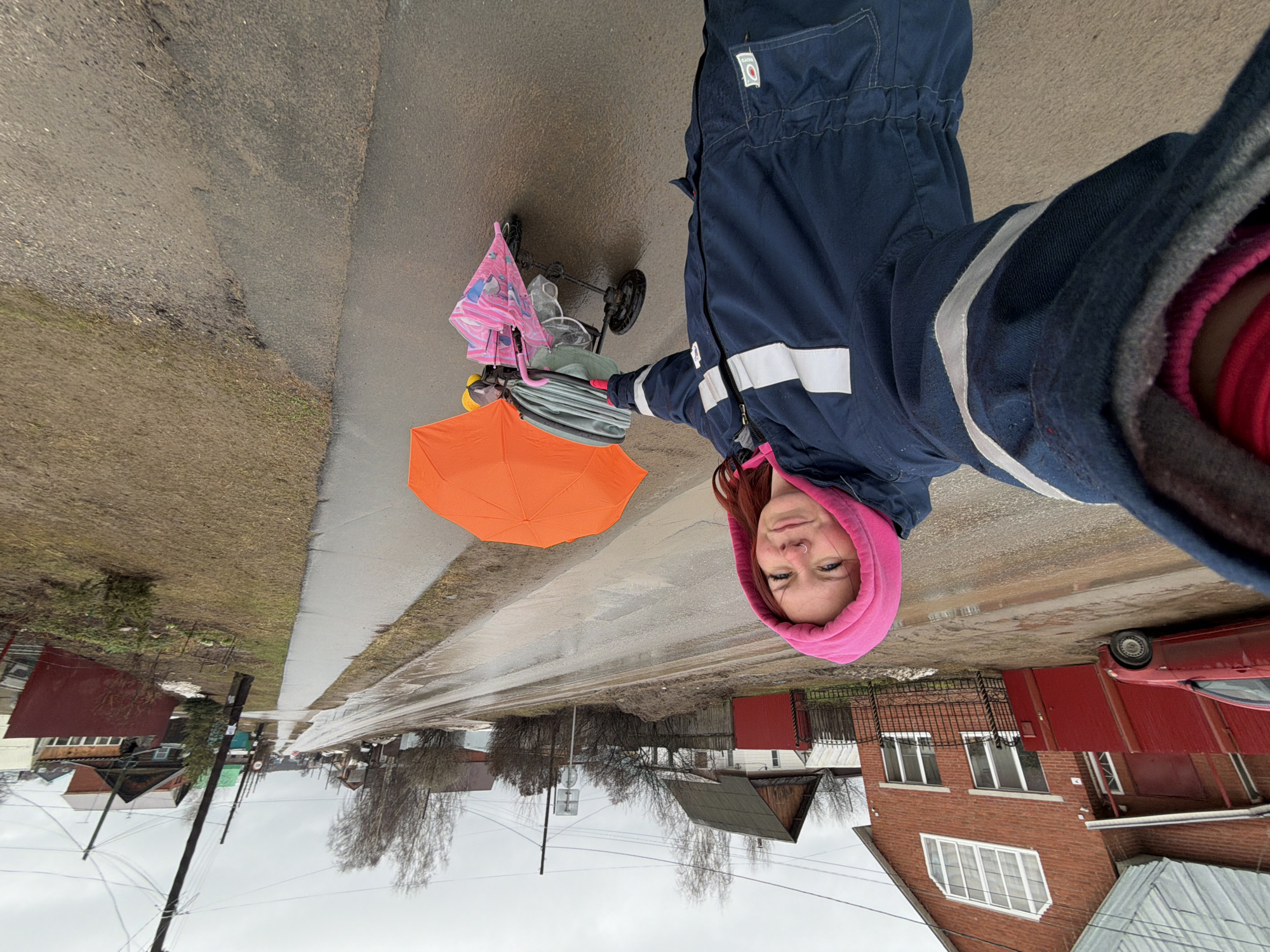 A selfie shows a woman on a wet, narrow street in a residential area, likely in Chaikovsky, Russia. The woman is wearing a blue coverall with white reflective strips and a pink hooded sweatshirt. She is holding a grey umbrella, and an open orange umbrella is positioned nearby. A baby stroller with a pink bag is visible to her right. The street appears recently wet, with reflections indicating pooled water. Houses with red brick or siding are visible on either side of the street, indicating a residential neighborhood. Electrical poles and bare trees are also present along the street. The sky is overcast. A red car is partially visible in front of one of the houses on the right side of the image.