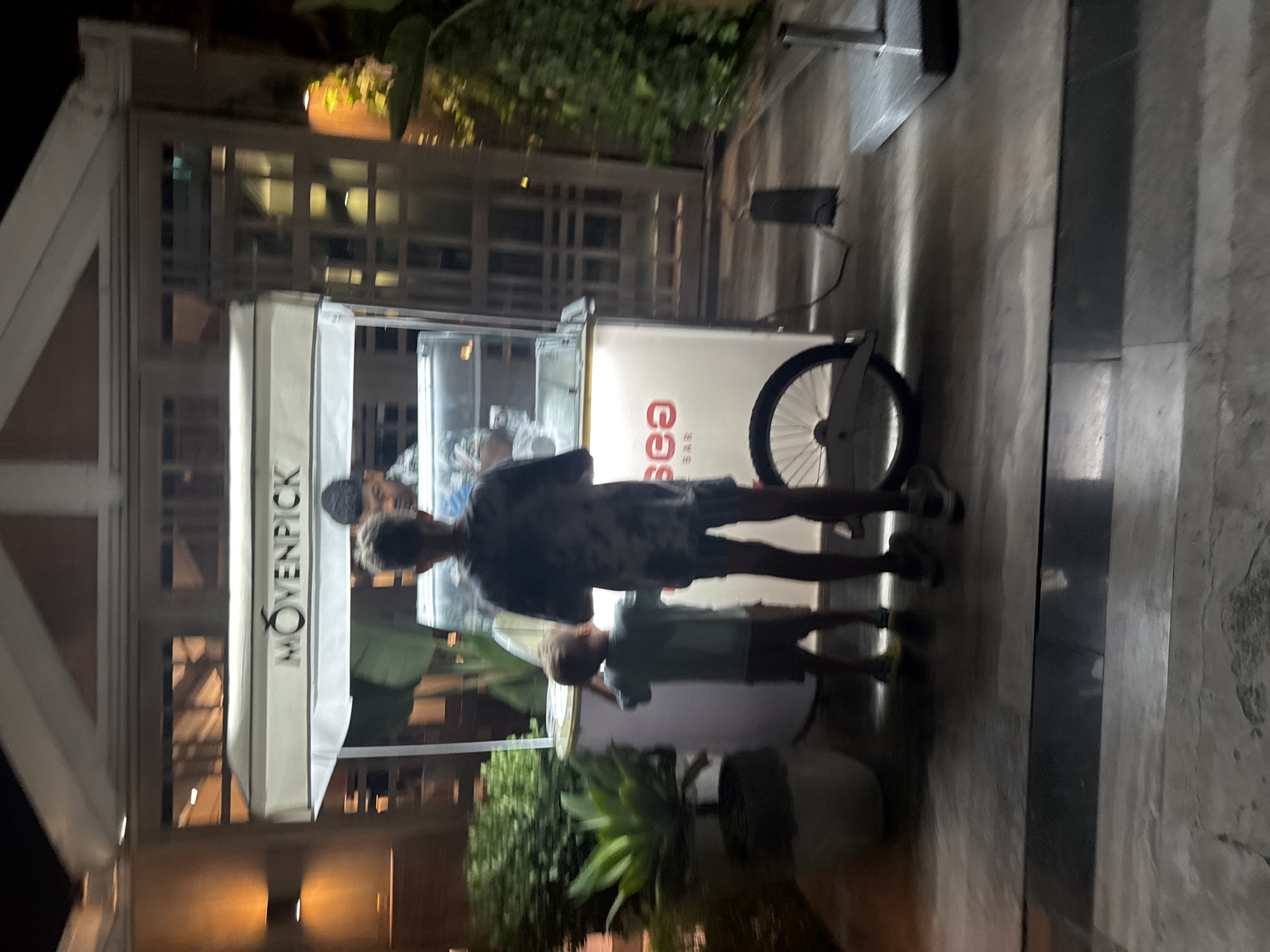 In front of a white and beige striped awning displaying the logo "MOVENPICK," an ice cream cart stands on a paved surface at night. A person, likely an employee, is visible behind the cart. Two children are in front of the cart, one looking towards the cart, and the other with their back to the camera. The cart has a glass display case and a white body with red lettering that reads "SEA BAR." A bicycle wheel is attached to the side of the cart. Green foliage is present to the left of the cart, and a building with windows and outdoor lighting is visible in the background. The scene appears to be located in a public area, possibly an outdoor dining or recreational space within the city of Parekklisia Community, Cyprus, given the context.