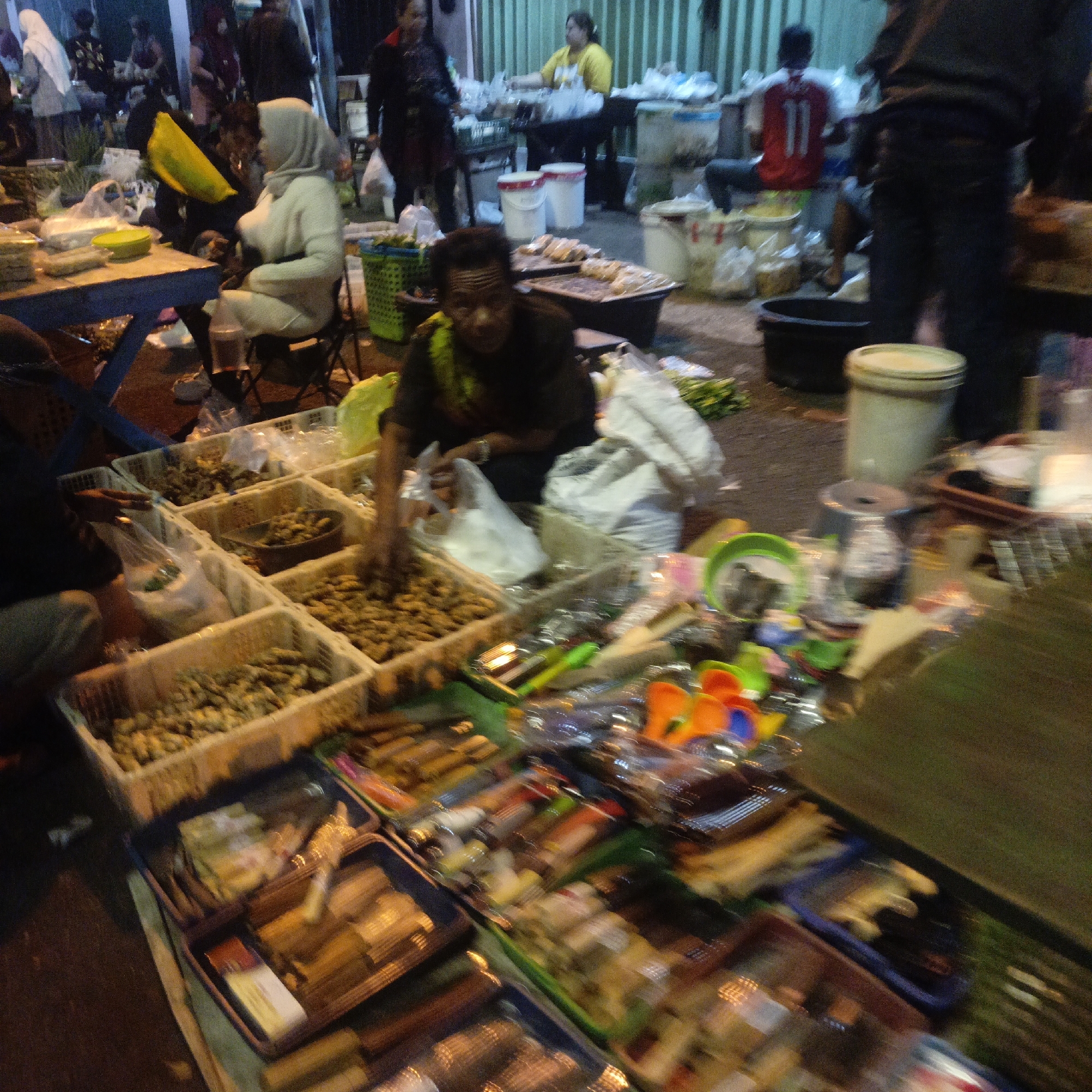 The image captures a bustling street market scene at dusk in Salatiga, Indonesia. In the foreground, a man wearing a dark shirt and a yellow sash is tending to a stall filled with an assortment of goods. He appears to be bagging some items. The stall itself is a collection of white plastic crates containing what look like dried goods, possibly snacks or ingredients, as well as packaged items. To the left, a blue table holds more items, and a yellow bowl is visible.
Behind the man, a woman wearing a white hijab and a light-colored sweater is seated on a small chair, seemingly observing the market. Further in the background, other individuals are present, some milling about and others attending to stalls. The market is set up along a paved street, with various stalls displaying goods in crates and on tables. A green corrugated metal wall is visible in the background, suggesting the market is set up against a building or structure. The overall atmosphere is one of lively commerce, with goods laid out for sale under the fading light.