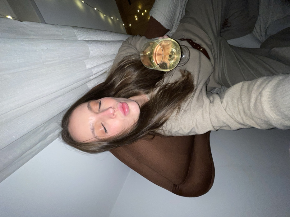 A young woman with light skin and long brown hair lies in a reclined position. She is holding a glass of white wine. She wears a beige sweater. She reclines on a brown chair with her back to a white curtain decorated with fairy lights.