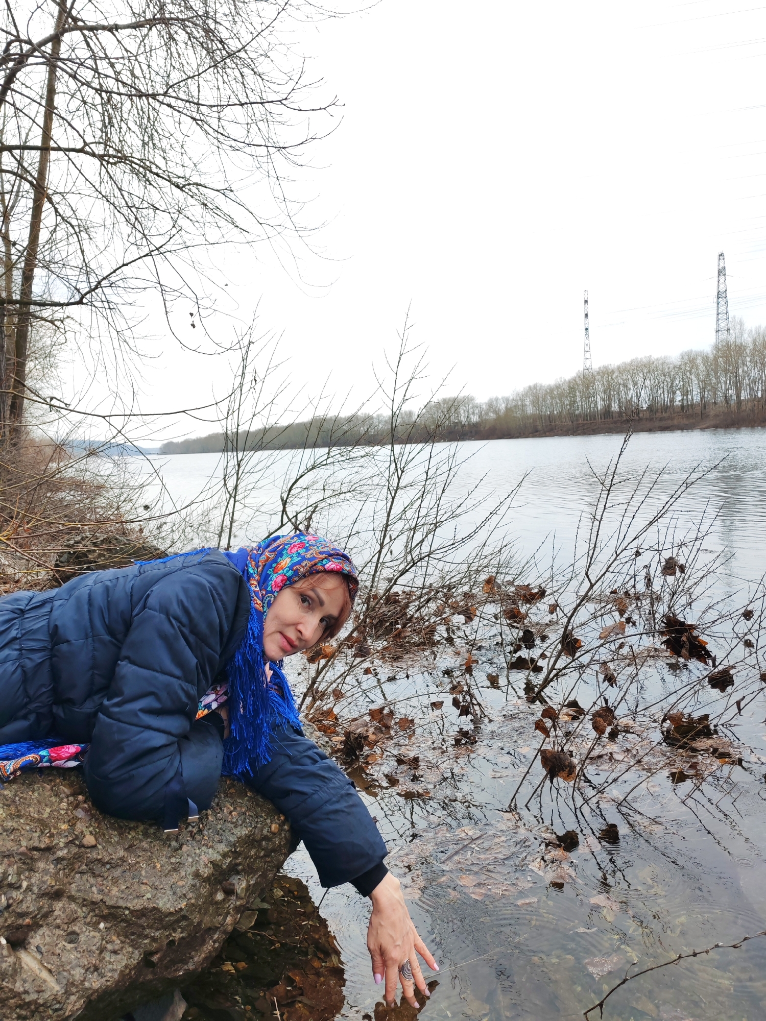 A woman is at the edge of a river. She is lying on a large rock at the river's edge and reaching her hand into the water. The woman is wearing a dark blue puffer jacket and a colorful scarf on her head. The river is wide and calm, and there are trees on the opposite bank. Power line towers are visible on the horizon. Bare tree branches and dead leaves are scattered around the water's edge. The sky is overcast. This appears to be on the outskirts of the city of Chaikovsky, Russia, where rivers often border wooded areas.