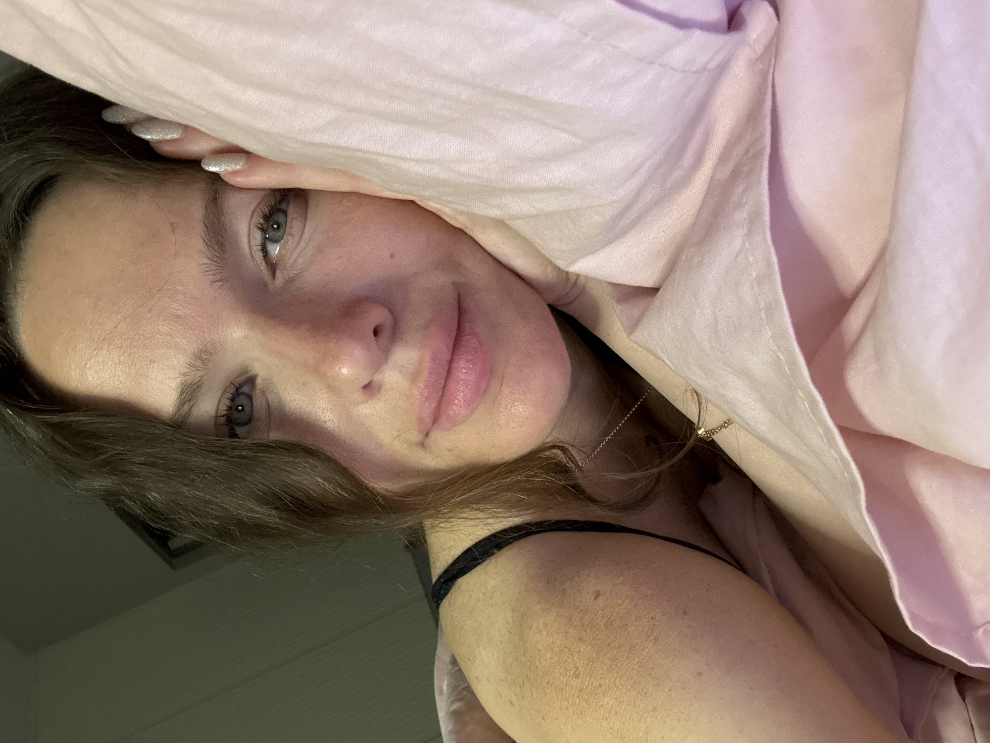 A close-up, eye-level, indoor, waist-up shot shows a woman lying down on pink bedding. The woman has fair skin with freckles, long brown hair, green eyes, and is wearing a black bra strap. She is resting her head on her hand, which has glittery silver nails. The background is dark and out of focus. No location in Khimki, Russia is depicted.