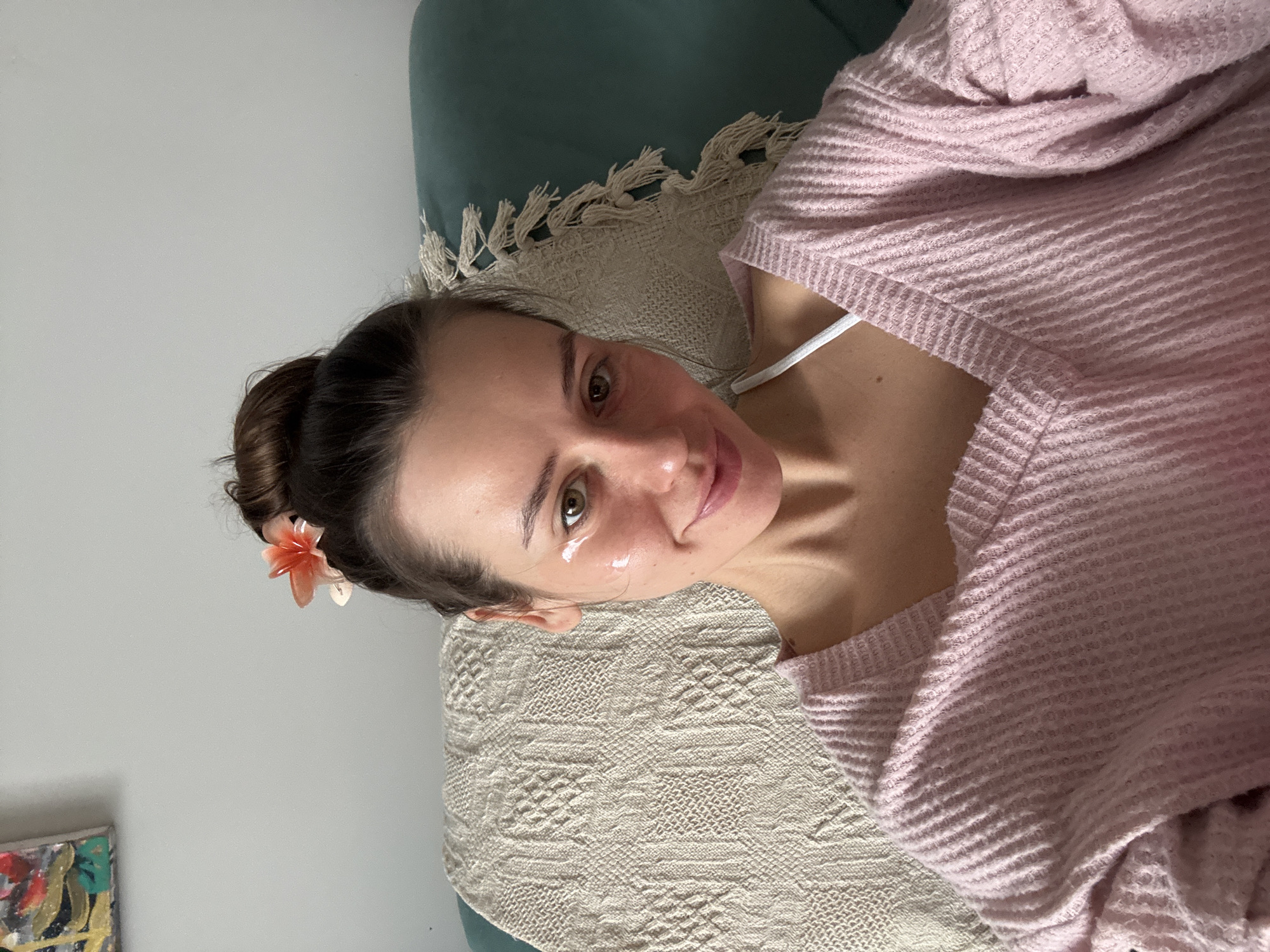 A young woman is captured in a close-up, head-and-shoulders portrait, lying down in what appears to be an indoor setting in Krasnoyarsk, Russia. She looks directly at the camera with a soft, inviting smile, her light brown or hazel eyes open wide. Her dark hair is styled in an updo or bun, with a delicate light orange/peach flower tucked into it on her left side.
She is wearing a light pink, textured, long-sleeved top, possibly a waffle-knit or ribbed sweater. A white strap, likely from an undershirt or bra, is visible on her right shoulder. Two distinct, glossy white droplets are visible under her right eye, suggesting the application of an eye cream or serum, indicating a moment of self-care or relaxation.
She is resting on a light-colored, possibly cream or beige, patterned blanket or throw, which features a woven geometric design and tassels. This blanket covers what appears to be a bed or sofa. Behind her head, a teal-colored upholstered pillow or headboard is visible. The background consists of a plain light gray wall, with a small section of an abstract painting featuring yellow, red, green, and black visible in the lower left corner. The lighting is soft and even, suggesting a bright daytime environment. The overall mood conveyed is calm and pleasant. There is no visible text in the image.
